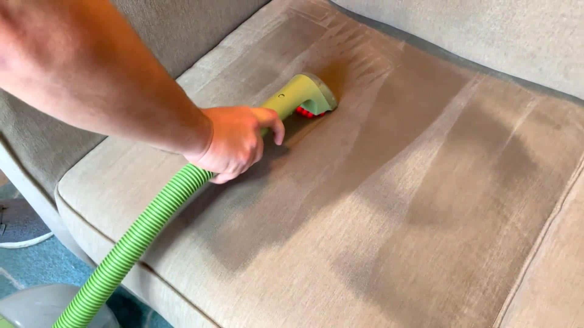 Cleaning 桌布 壁紙 — 一隻手引導綠色蒸氣清潔器在米色布藝沙發墊上移動，顯示出乾淨條紋與骯髒部分的對比。