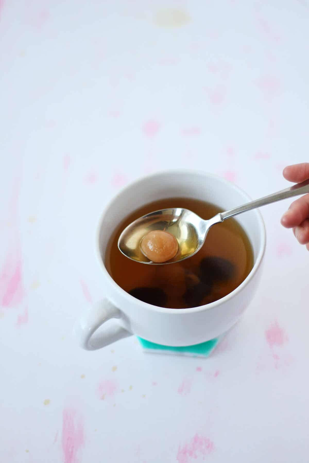 茶 桌布 壁紙 — 一隻手從一個裝有琥珀色液體的白色杯子中提起一個湯匙，湯匙上有一顆半透明的棕色珍珠，背景是柔和的粉紅色潑濺。