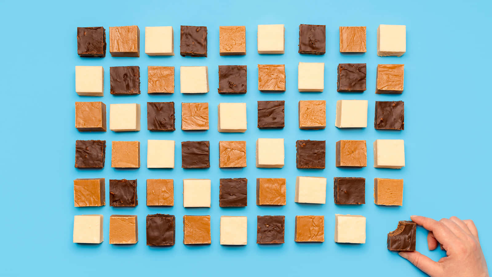 Penuche Fudge 桌布 壁紙 — 一隻手從亮藍色背景上各種軟糖方塊的網格中揀選一塊巧克力軟糖方塊。