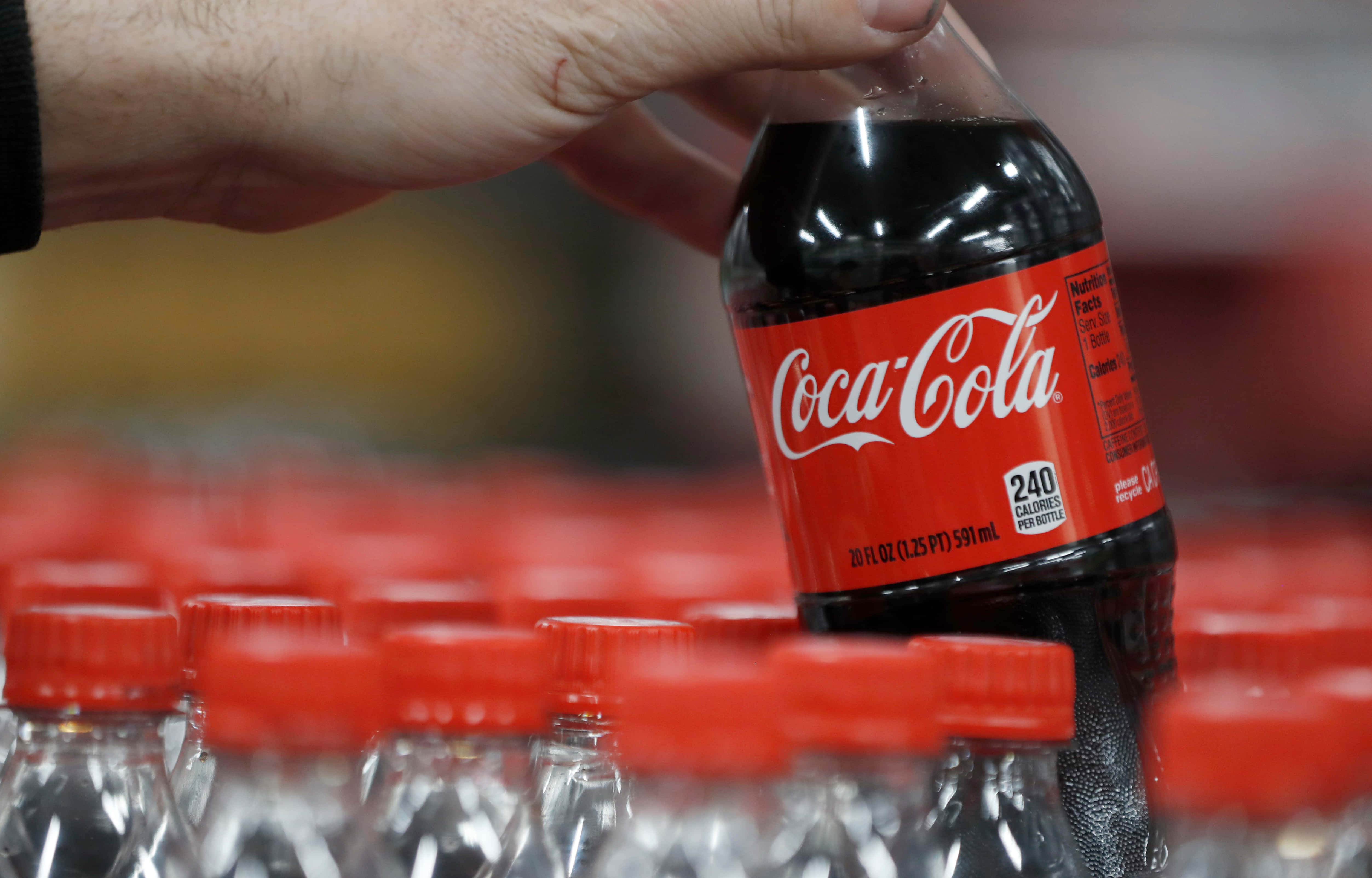 coca cola 桌布 壁紙 — 一隻手從商店貨架上拿起一瓶冰涼的可口可樂，貨架上擺滿了相同的瓶子。