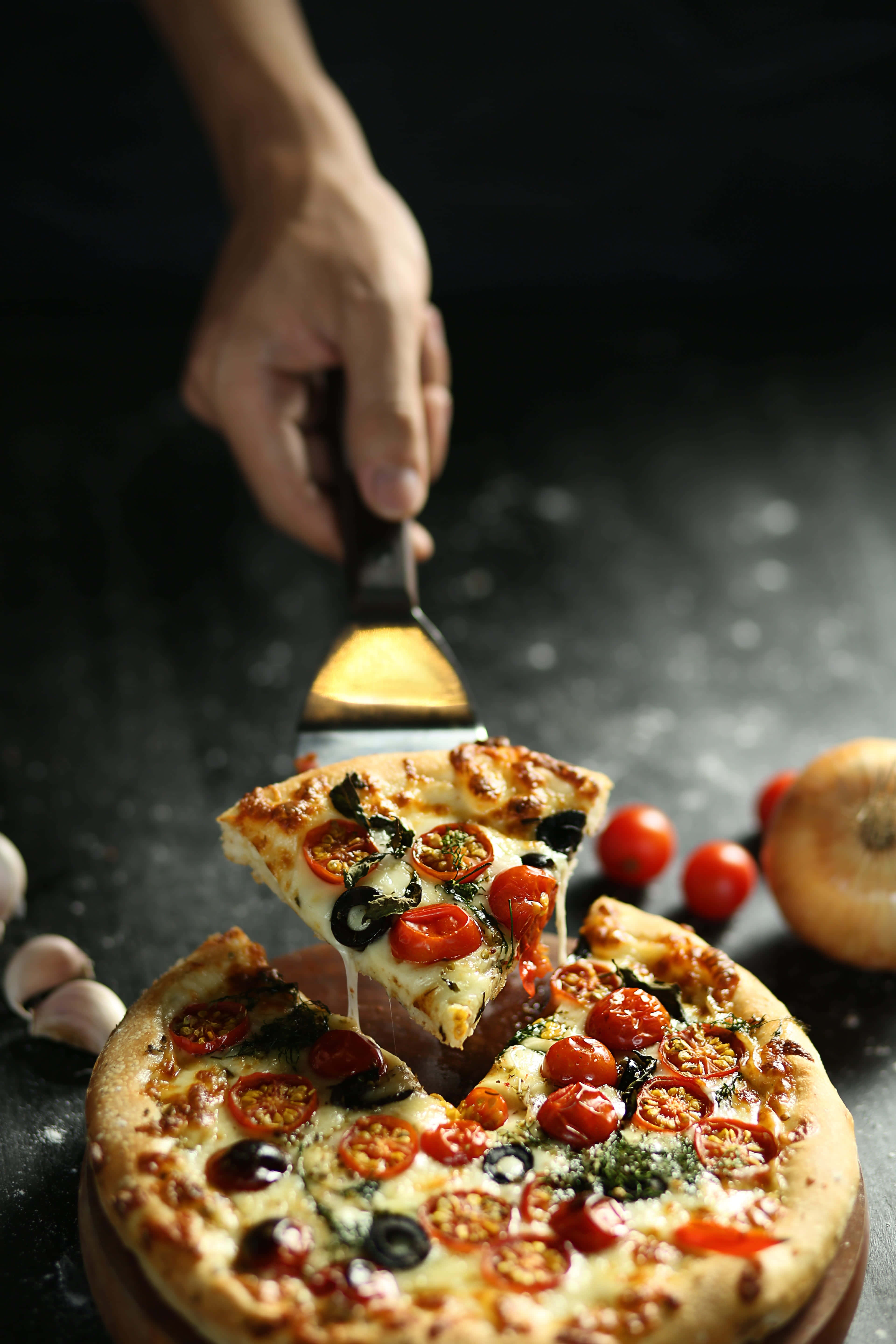 Pizza 桌布 壁紙 — 一隻手從深色鄉村風格的表面拿起一片鋪滿小番茄和橄欖的起司披薩。