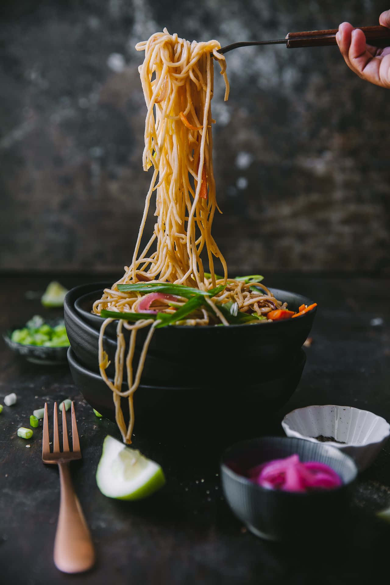 Food Photography 桌布 壁紙 — 一隻手從黑色碗中提起冒煙的麵條，周圍是鮮豔的裝飾和調味料，放在深色鄉村風格的表面上。