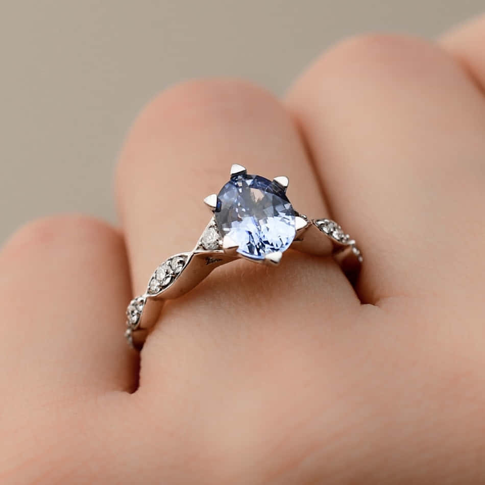 rings 桌布 壁紙 — 一隻手戴著一枚令人驚嘆的藍寶石訂婚戒指，周圍鑲嵌著鑽石光環，採用銀色金屬材質。