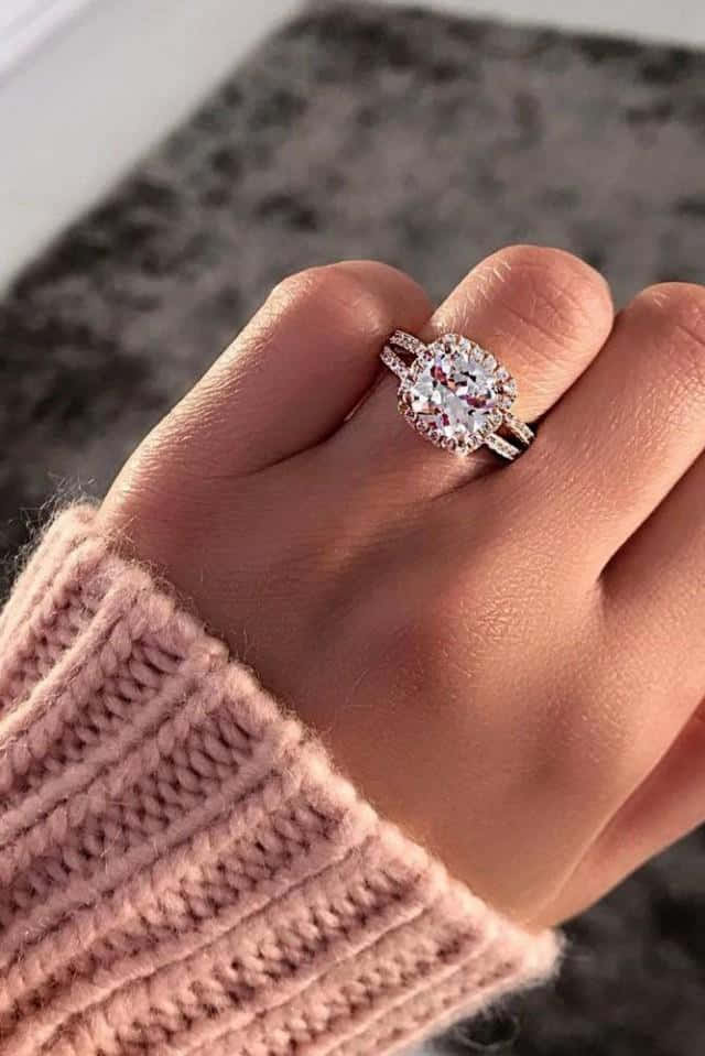 rings 桌布 壁紙 — 一隻手戴著閃閃發光的光環訂婚戒指，配粉紅色針織袖，背景是柔和的灰色紋理。