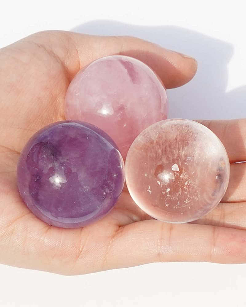水晶 桌布 壁紙 — 一隻手托住三顆拋光寶石球體—紫水晶、玫瑰石英和透明石英—在白色背景的柔和光線下閃閃發光。