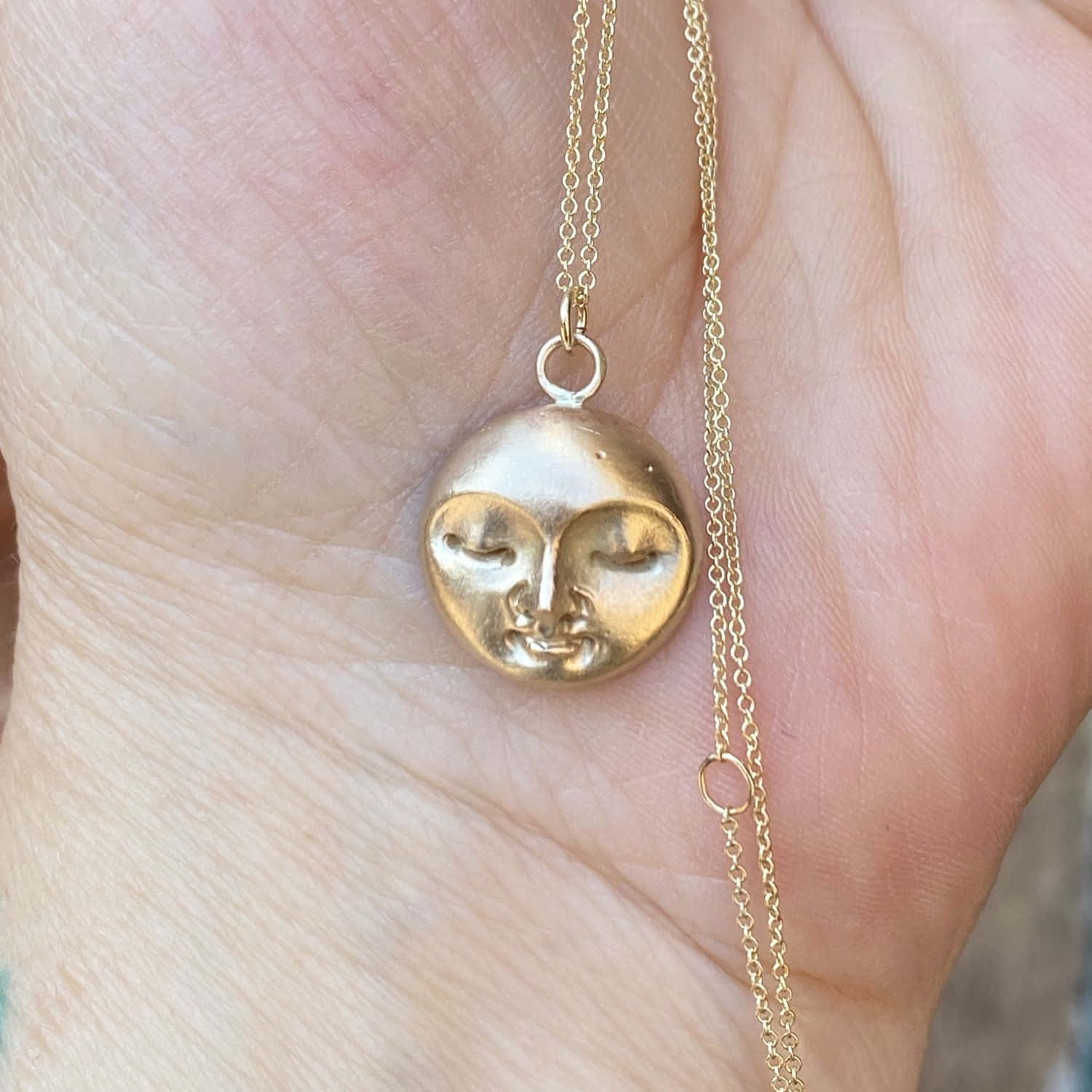 Moon Face 桌布 壁紙 — 一隻手托著一條精緻的金項鍊，上面有一個圓形吊墜，設計成一個寧靜、閉眼的臉孔。