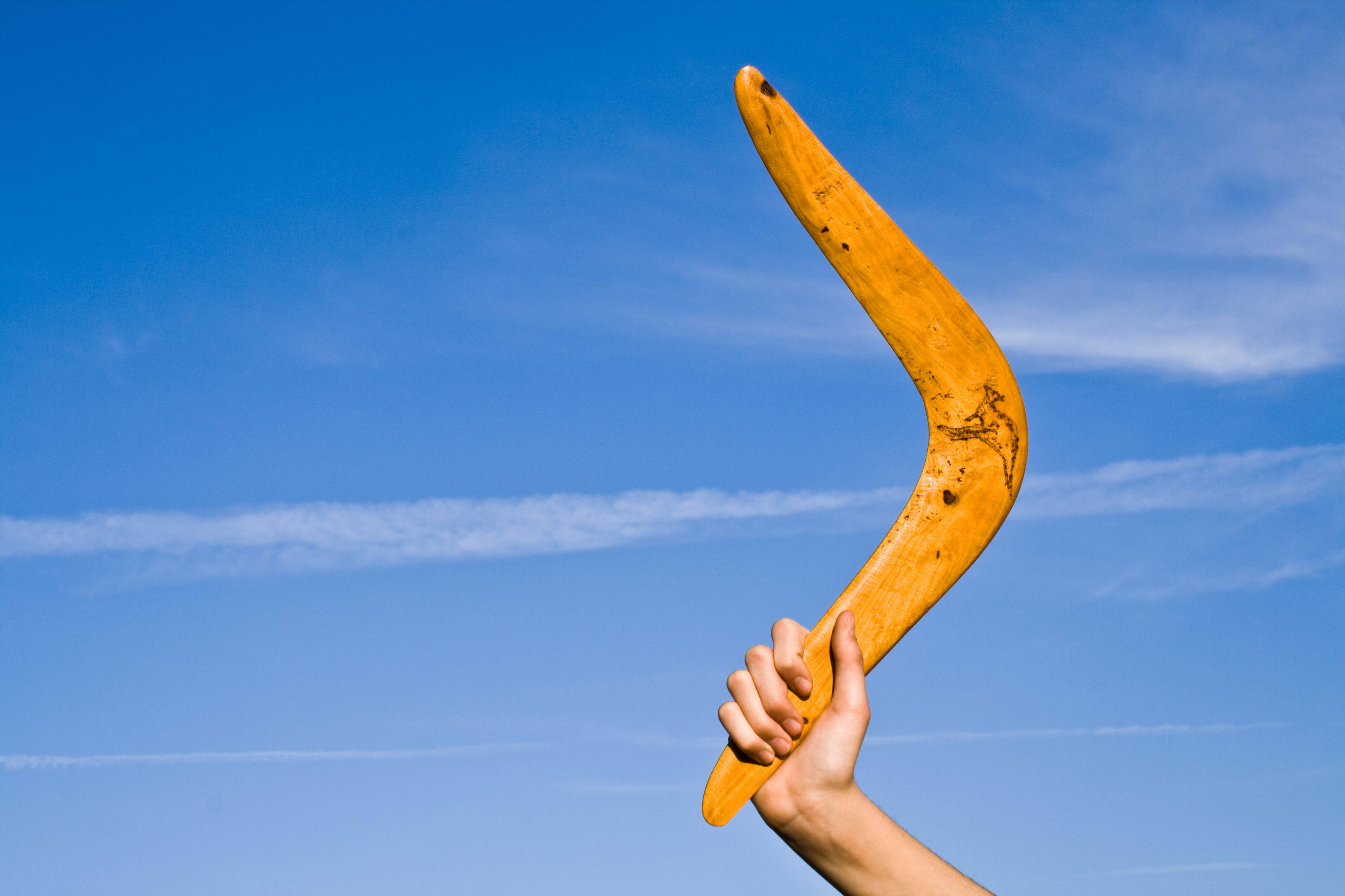 Boomerang 桌布 壁紙 — 一隻手抓住一個彎曲的木製迴力鏢，背景是晴朗的藍天和薄雲。