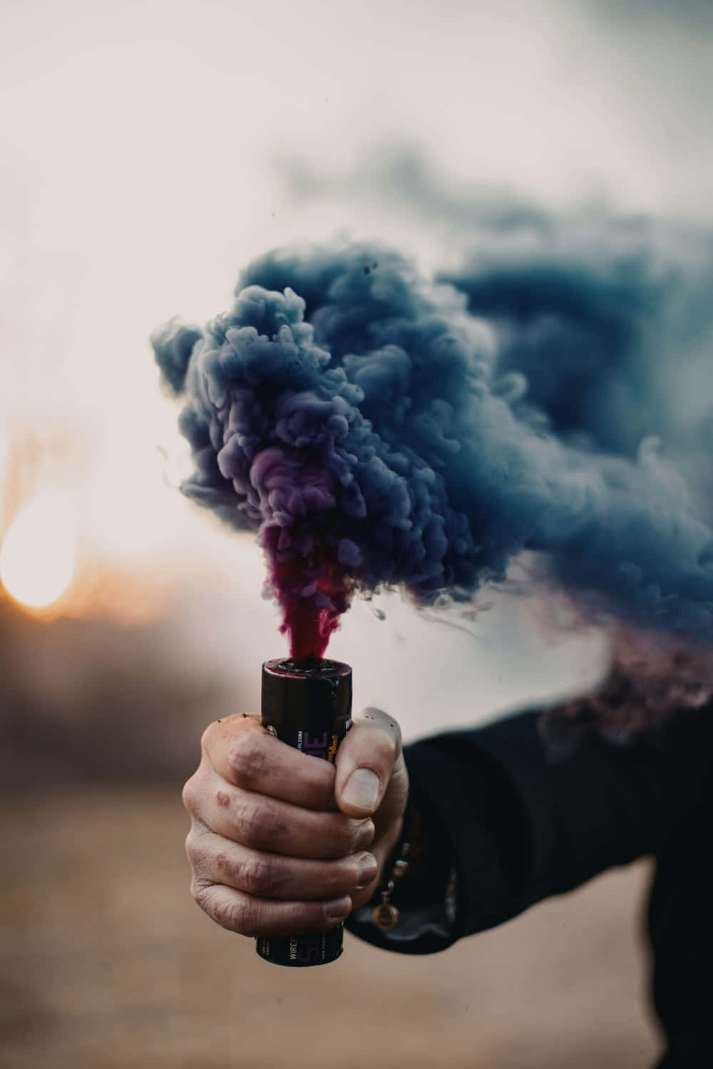桌布 壁紙 — 一隻手抓住一個煙霧彈，在柔和的日落背景下釋放出充滿活力的紫色和藍色煙霧。