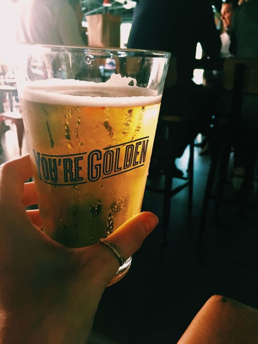 啤酒 桌布 壁紙 — 一隻手抓住一個結霜的啤酒杯，上面有「YOU'RE GOLDEN」字樣和泡沫，在燈光昏暗的酒吧裡。