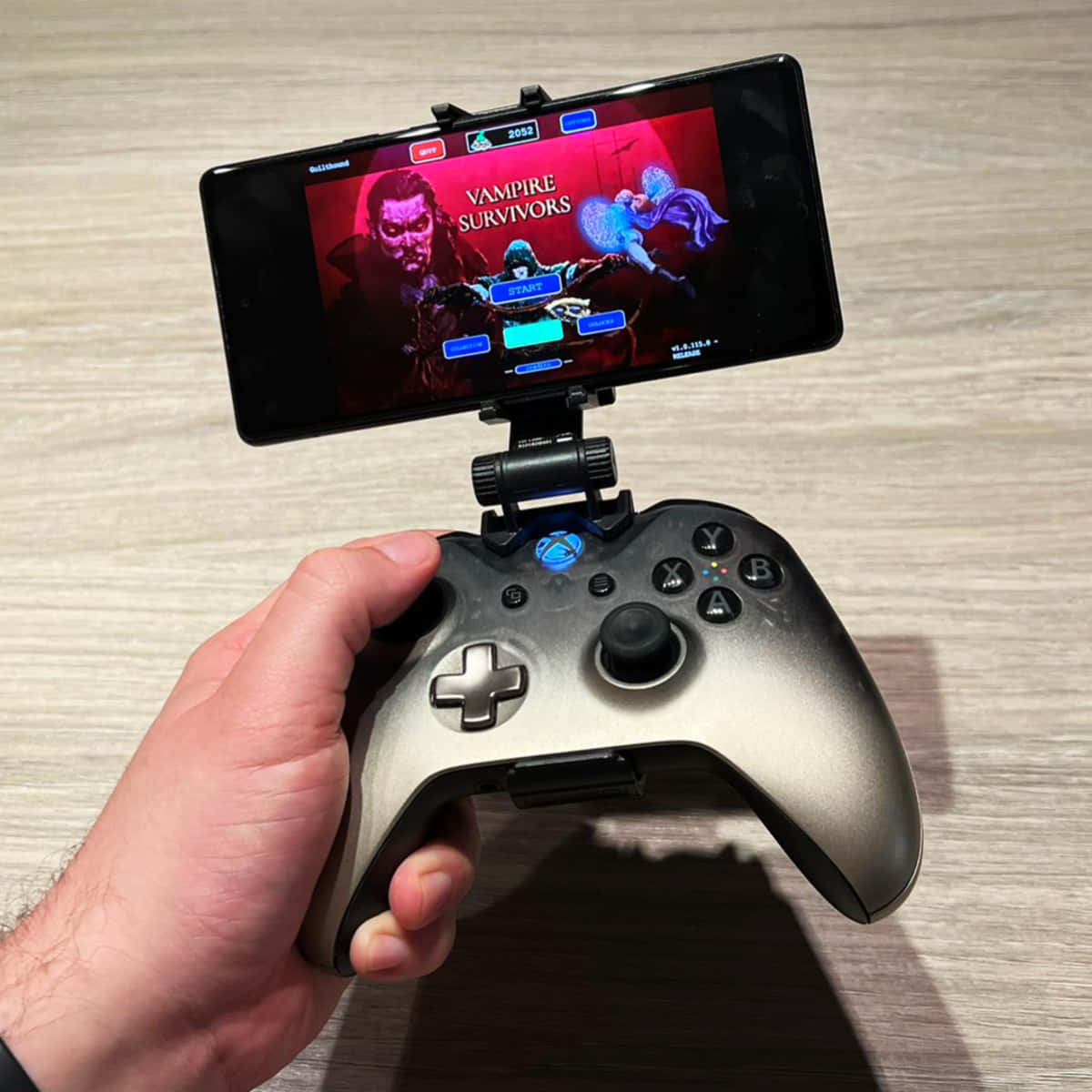 Xbox 桌布 壁紙 — 一隻手抓住一個銀色Xbox控制器，安裝在智慧型手機上玩Vampire Survivors，手機顯示一個黑暗奇幻遊戲選單。
