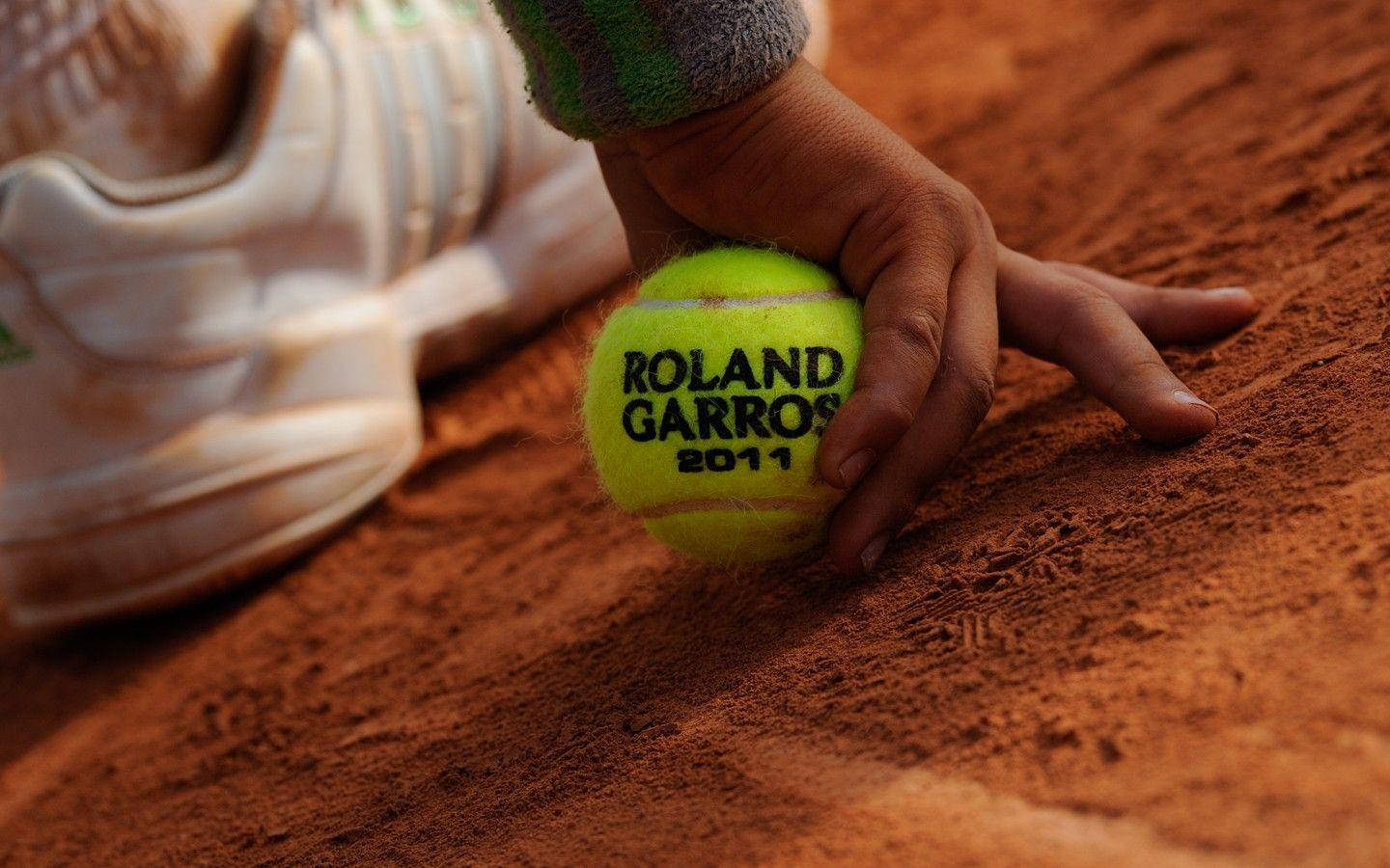 french 桌布 壁紙 — 一隻手抓住一個鮮黃色的Roland Garros 2011網球，在紋理紅土球場上，旁邊有一隻白色運動鞋。