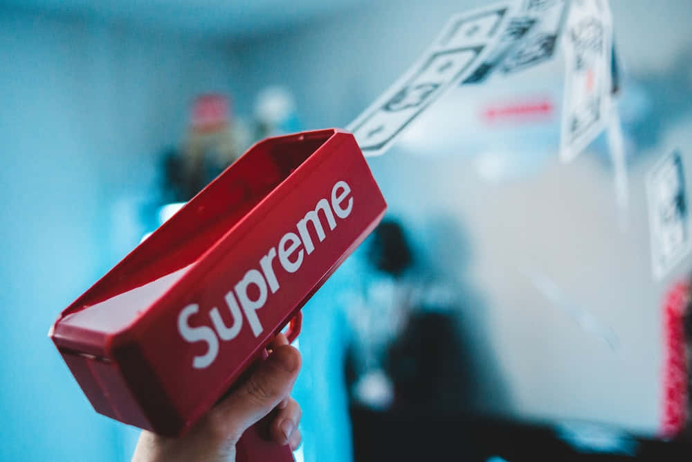 supreme 桌布 壁紙 — 一隻手抓住一把紅色的Supreme玩具槍，美元鈔票在模糊的藍色背景中飛舞。