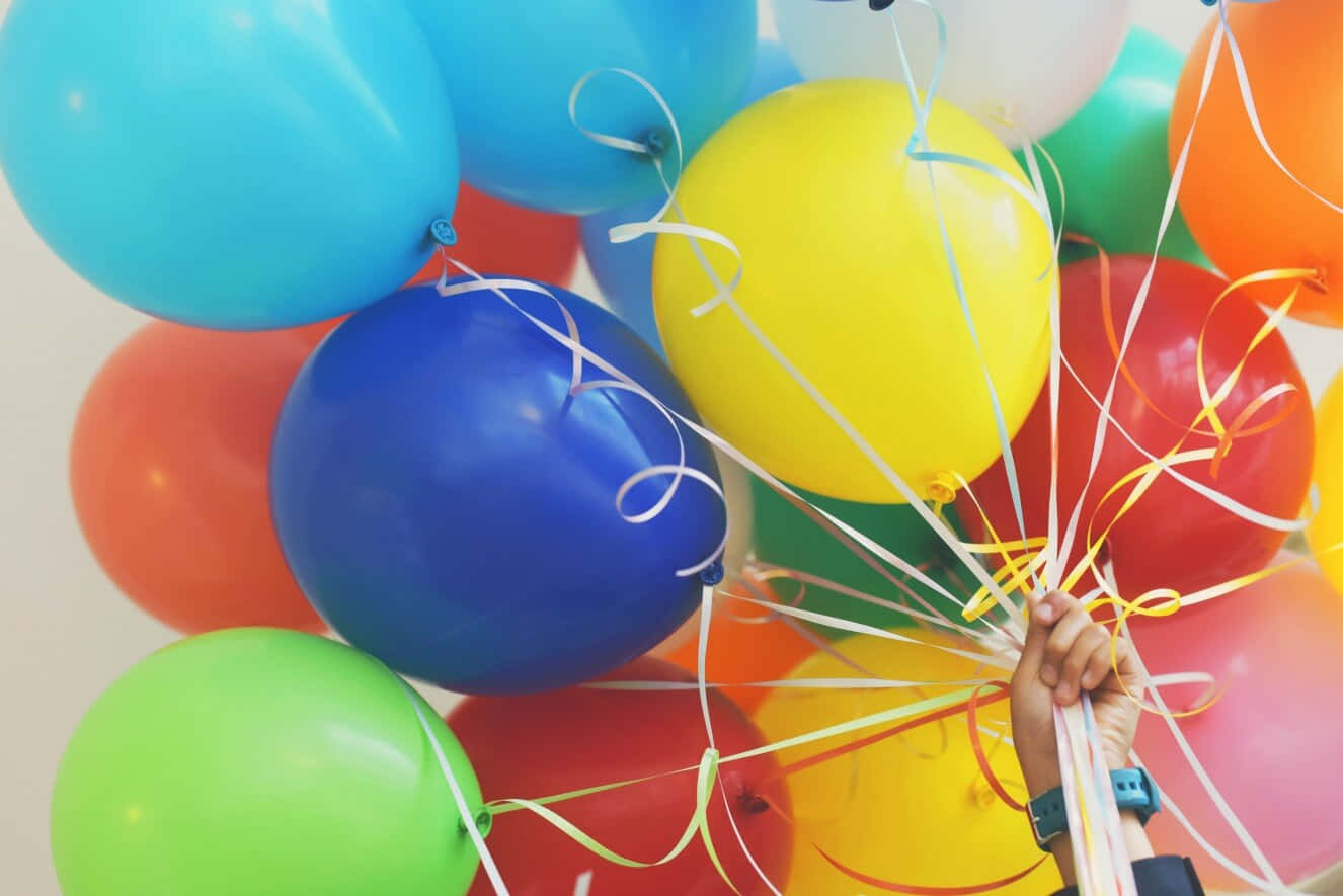 Birthday Party 桌布 壁紙 — 一隻手抓住一束鮮豔的氣球，顏色有紅、藍、黃、綠和橙色，絲帶在純色背景上飄揚。