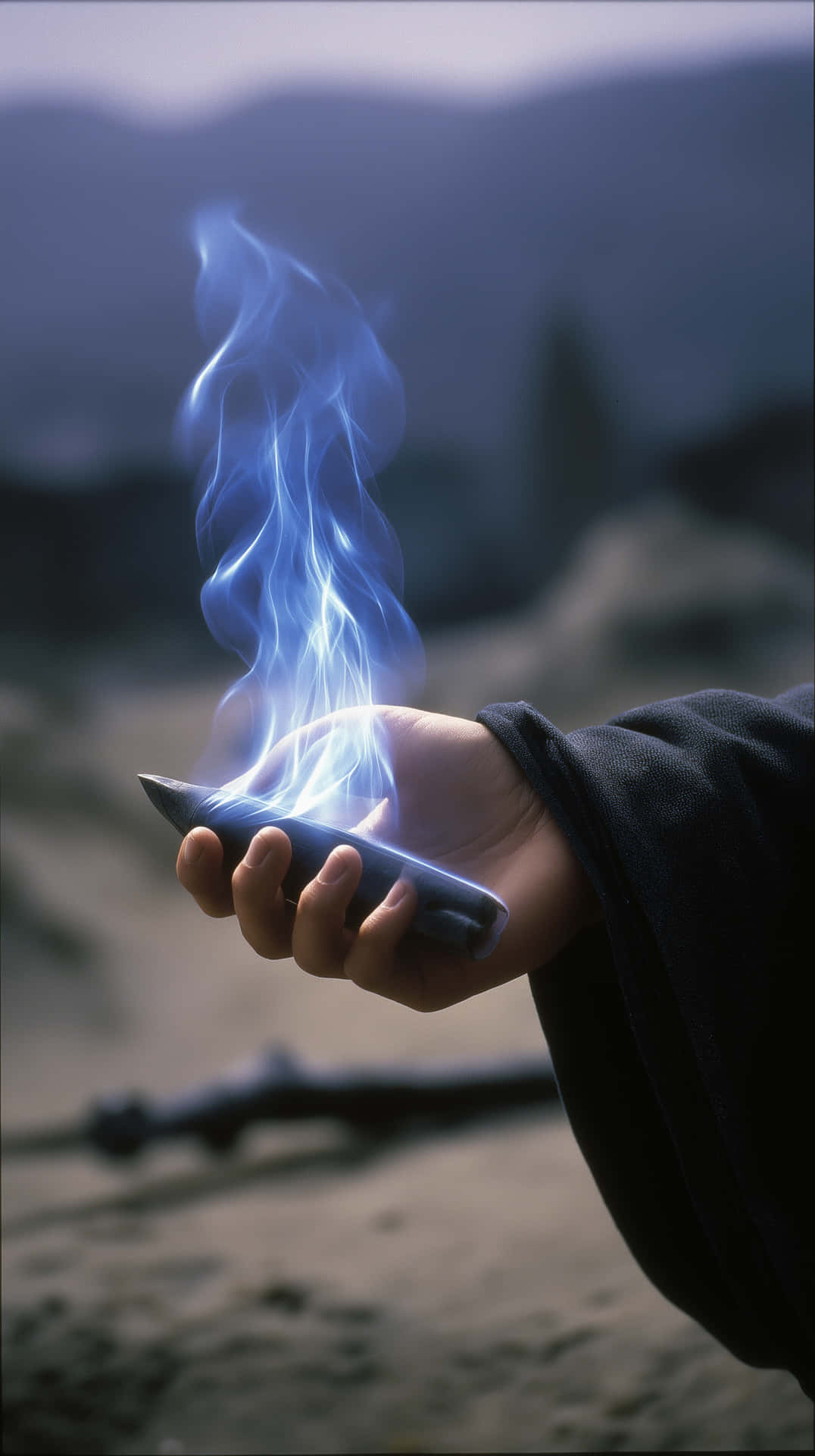 Hand Holding Object Emitting Blue Flames in Dark Setting 壁紙 — 一隻手抓住發光的藍色能量刀片，背景是模糊的山脈。