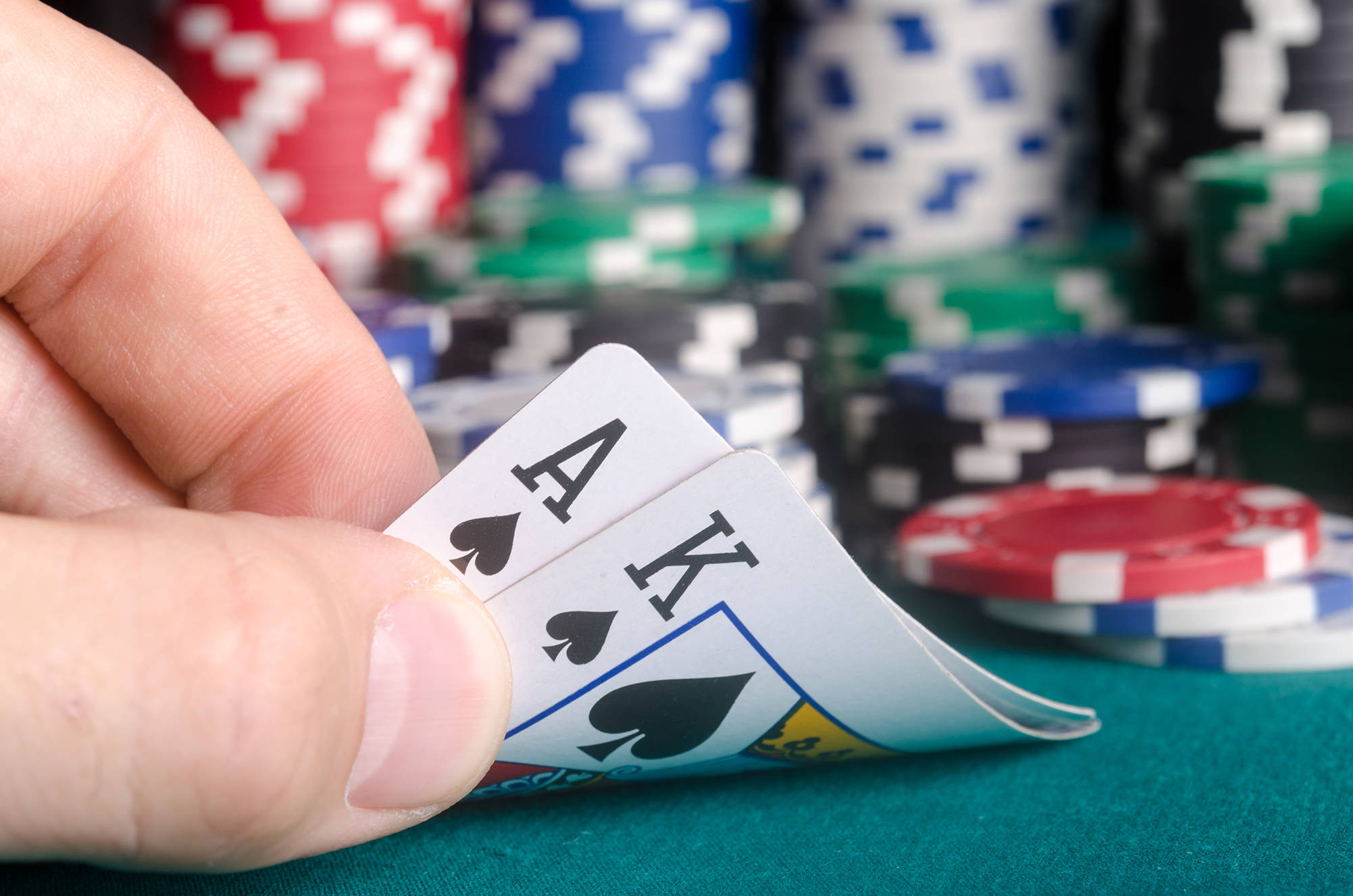Casino 桌布 壁紙 — 一隻手抓住黑桃A和K牌，上方是綠色絨布桌，背景有彩色籌碼堆疊。