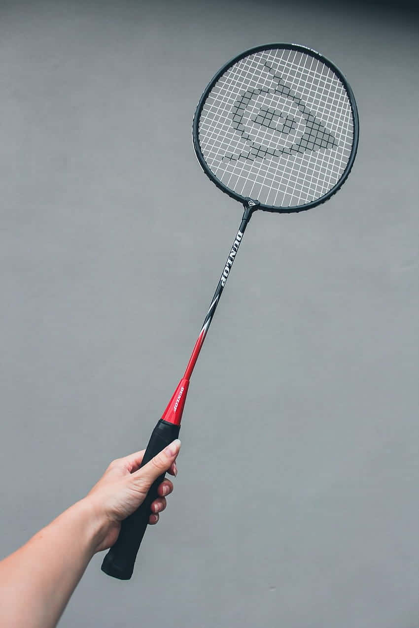 Android Badminton 桌布 壁紙 — 一隻手抓住黑色和紅色的Dunlop羽毛球拍，背景是純灰色工作室。