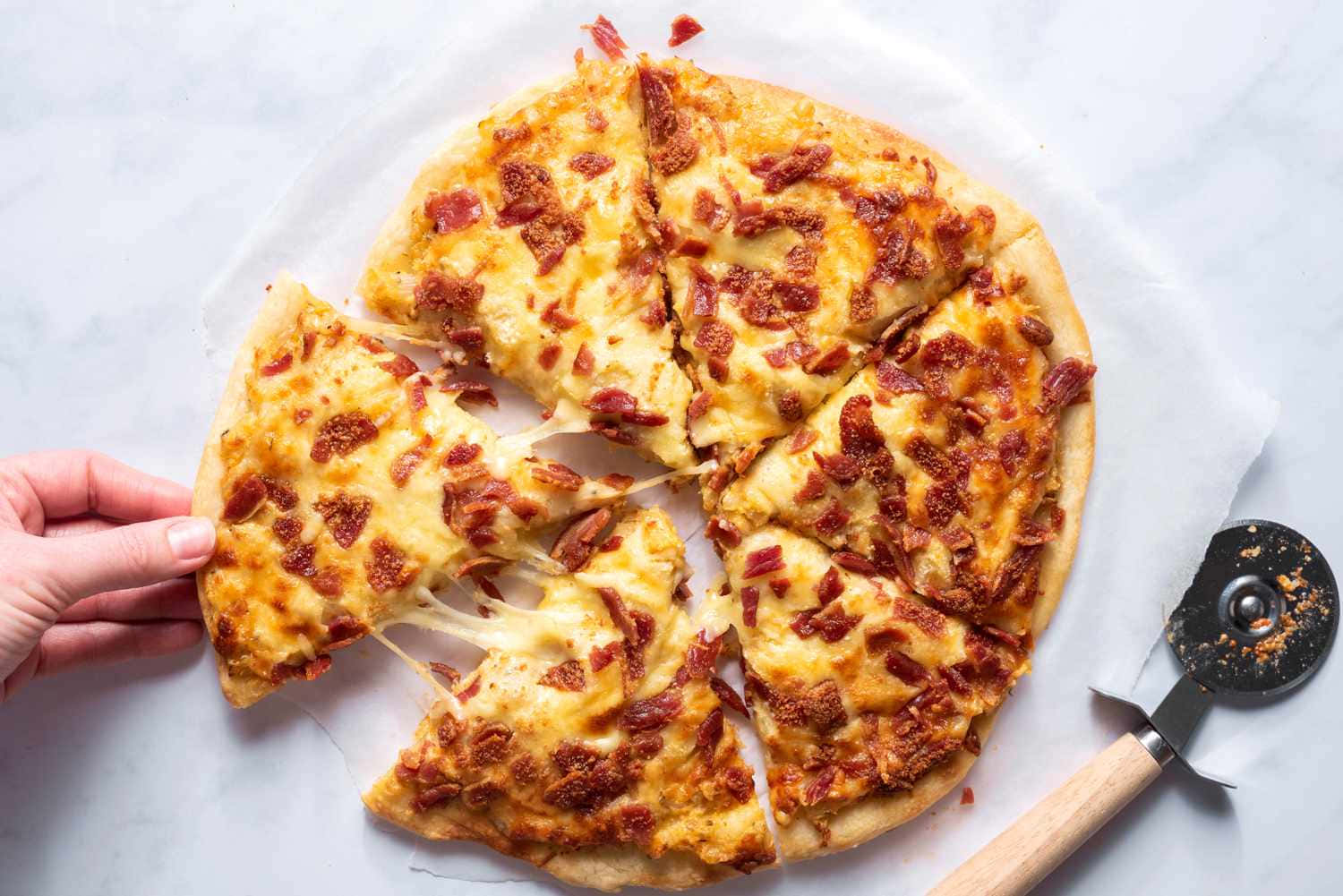 Pizza 桌布 壁紙 — 一隻手拉出一塊起司培根披薩，露出牽絲起司，背景是大理石，附近有一個披薩切刀。