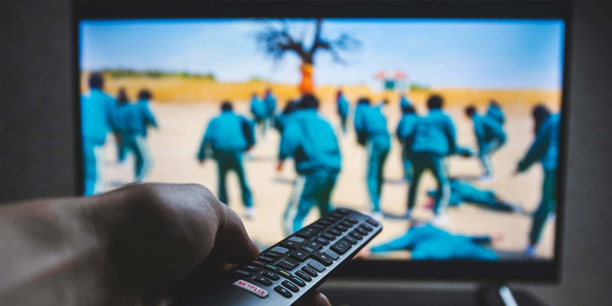 Squid Game 桌布 壁紙 — 一隻手拿著 Netflix 遙控器，對準電視，電視上正在播放一個緊張場景，節目中的人穿著藍色制服，身處荒蕪的景觀中。