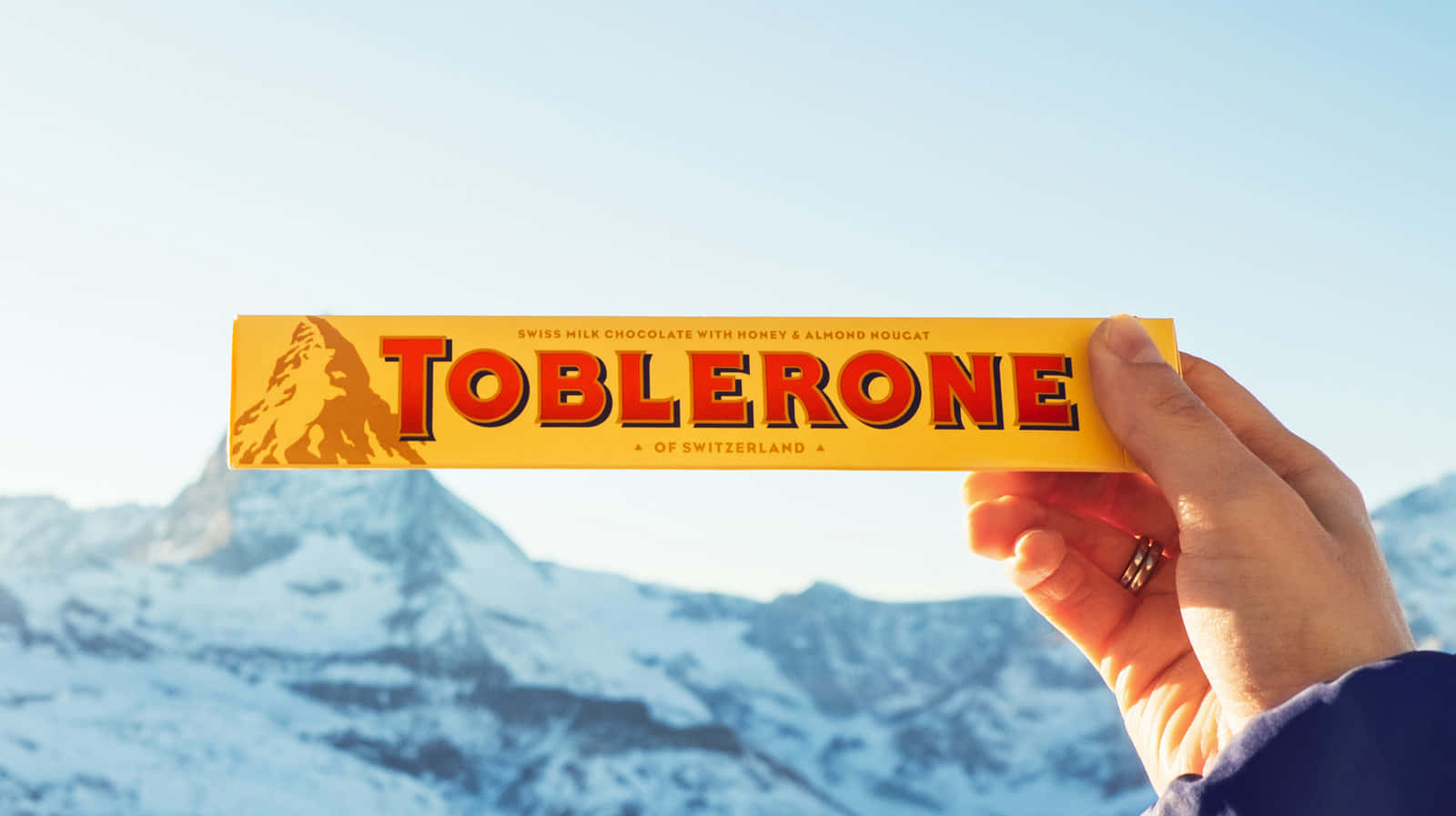 Chocolate 桌布 壁紙 — 一隻手拿著 Toblerone 巧克力棒，背景是晴朗藍天下白雪覆蓋的瑞士山脈。