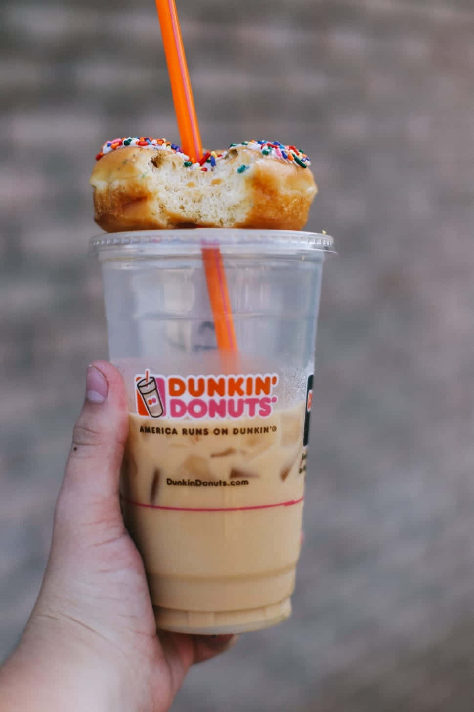 Iced Coffee 桌布 壁紙 — 一隻手拿著Dunkin' Donuts冰咖啡，上面放著半個吃剩的彩針甜甜圈和一根橙色吸管。