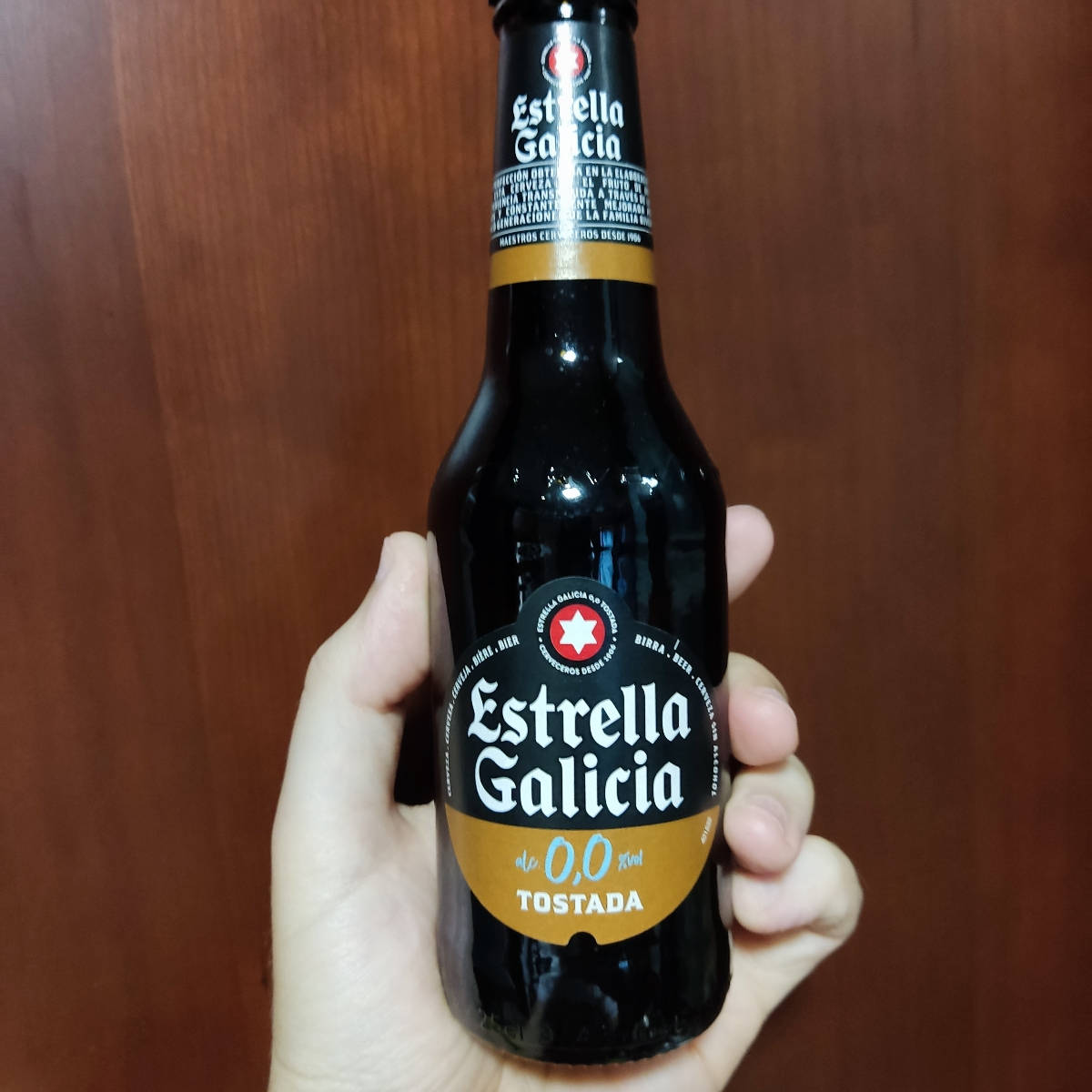 Beer 桌布 壁紙 — 一隻手拿著Estrella Galicia 0,0 Tostada啤酒的深色玻璃瓶，背景是溫暖的木質。