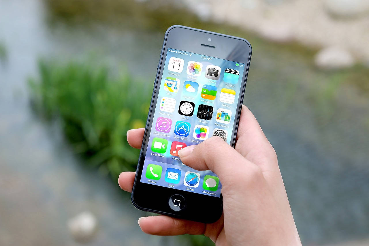 科技動圖 桌布 壁紙 — 一隻手拿著iPhone，在模糊的自然水景和綠植背景上顯示色彩繽紛的應用程式圖標。
