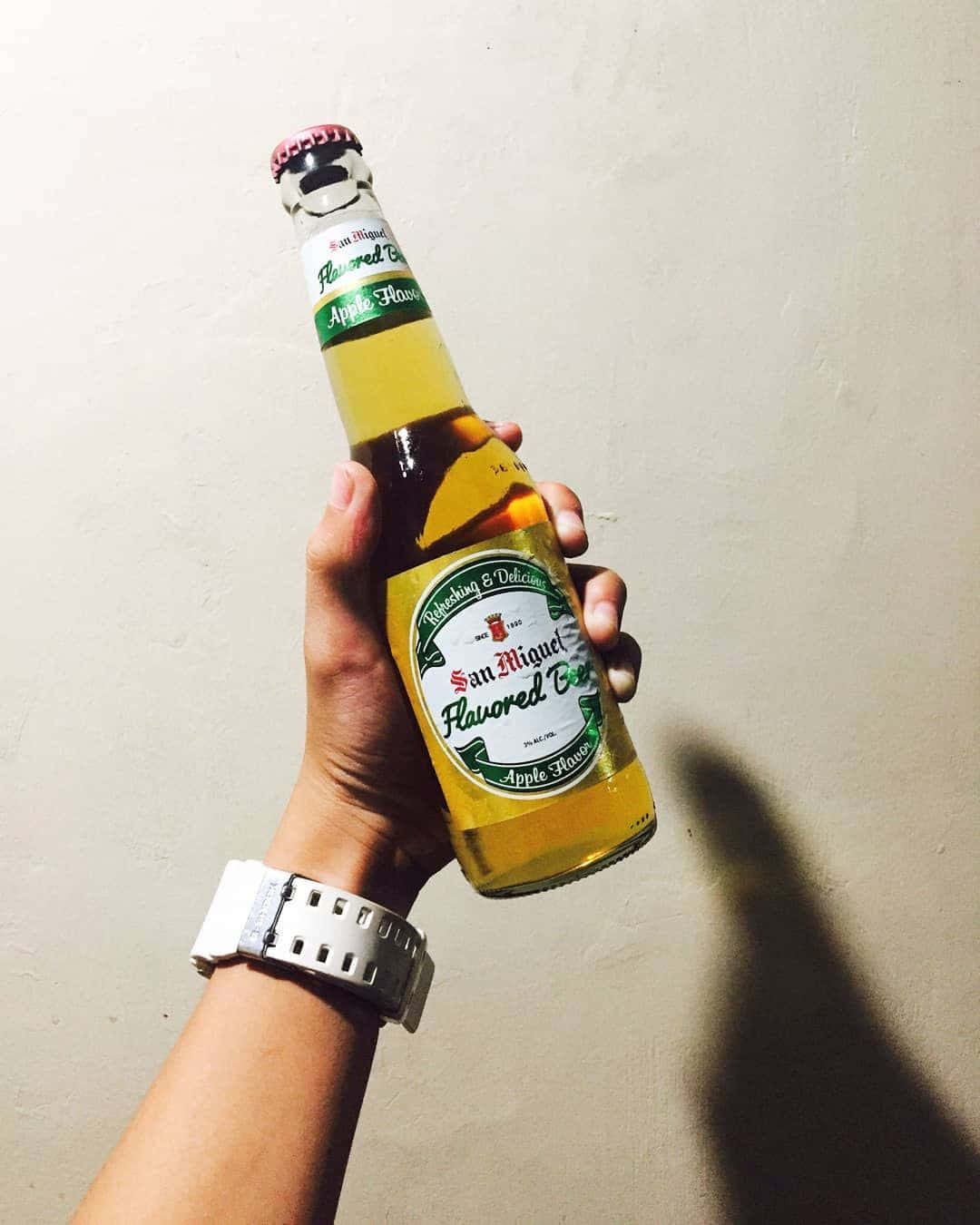 Beer 桌布 壁紙 — 一隻手拿著San Miguel蘋果味啤酒瓶靠著素色牆壁，手腕上戴著手錶。