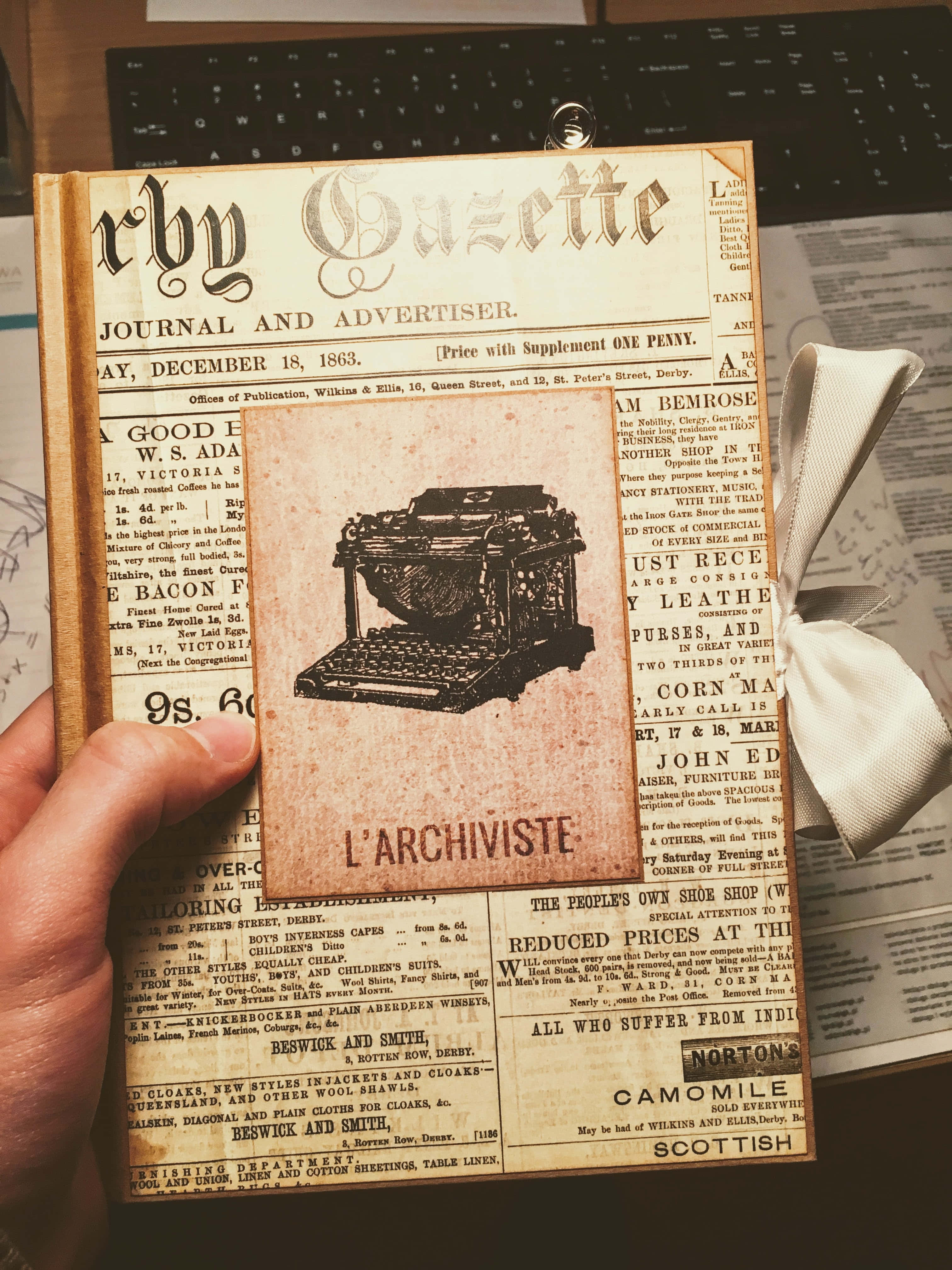 Old Newspaper 桌布 壁紙 — 一隻手拿著一份名為《Derby Gazette》的復古報紙，日期為1863年12月18日，上面有古董打字機插圖和褪色的廣告。