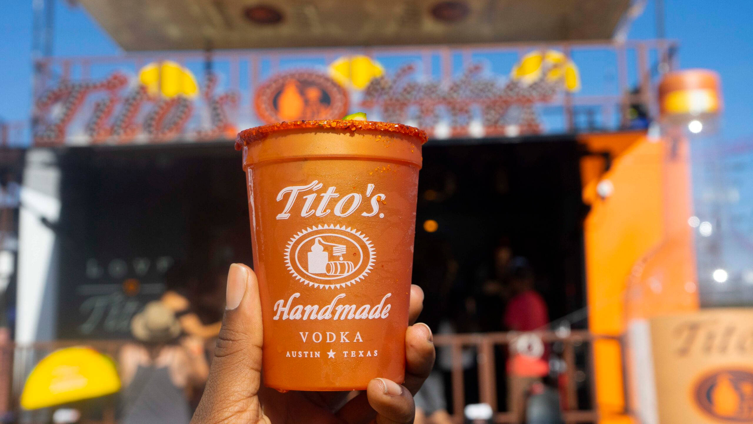 cup 桌布 壁紙 — 一隻手拿著一個充滿活力的橙色Tito's Handmade Vodka杯，杯緣沾有紅色香料，背景是陽光明媚的節日場景。
