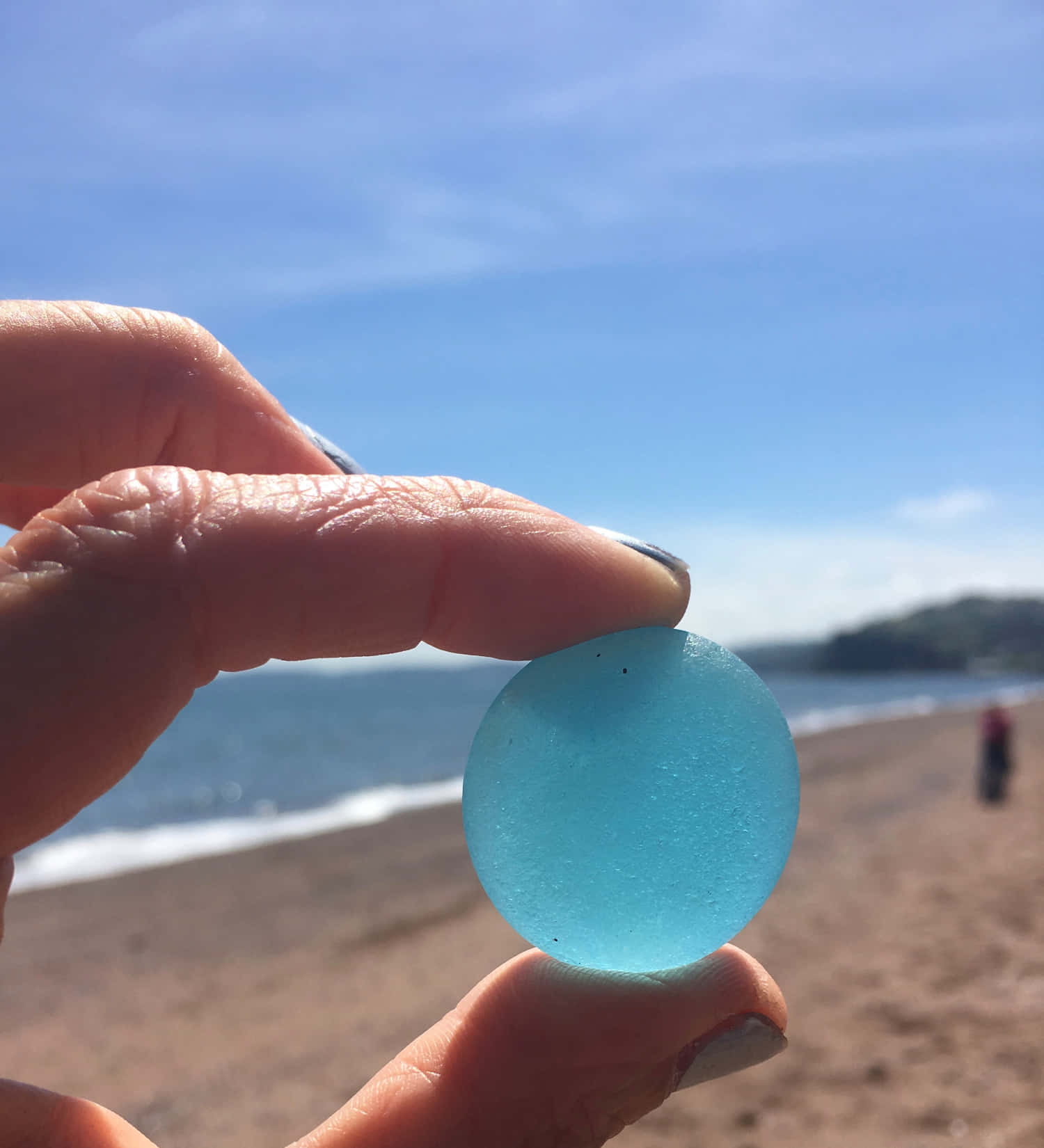 Sea Glass 桌布 壁紙 — 一隻手拿著一個光滑半透明的藍色海玻璃球體，背景是陽光明媚的海灘，有輕柔的海浪和遠處的山丘。