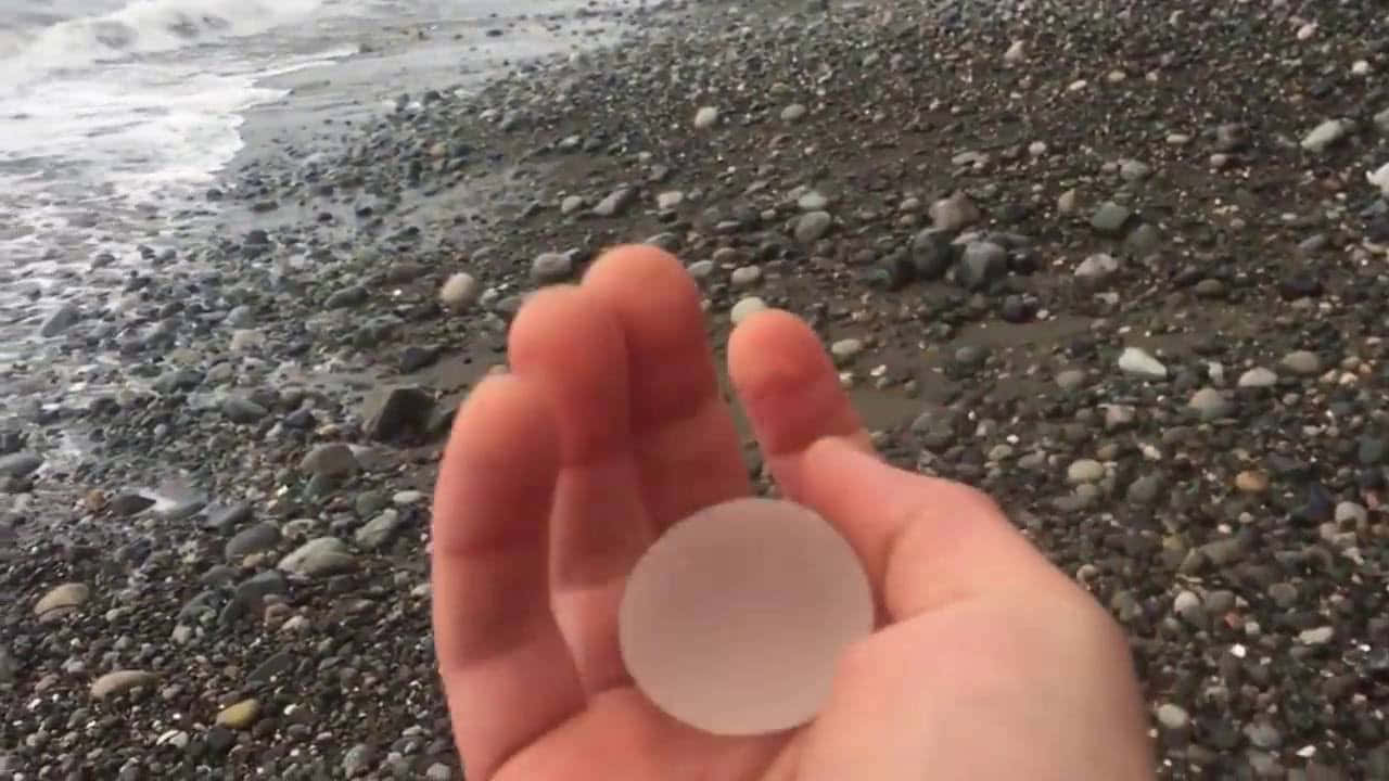 Sea Glass 桌布 壁紙 — 一隻手拿著一個光滑的磨砂玻璃海洋圓盤，背景是濕漉漉的鵝卵石和拍打的海浪。