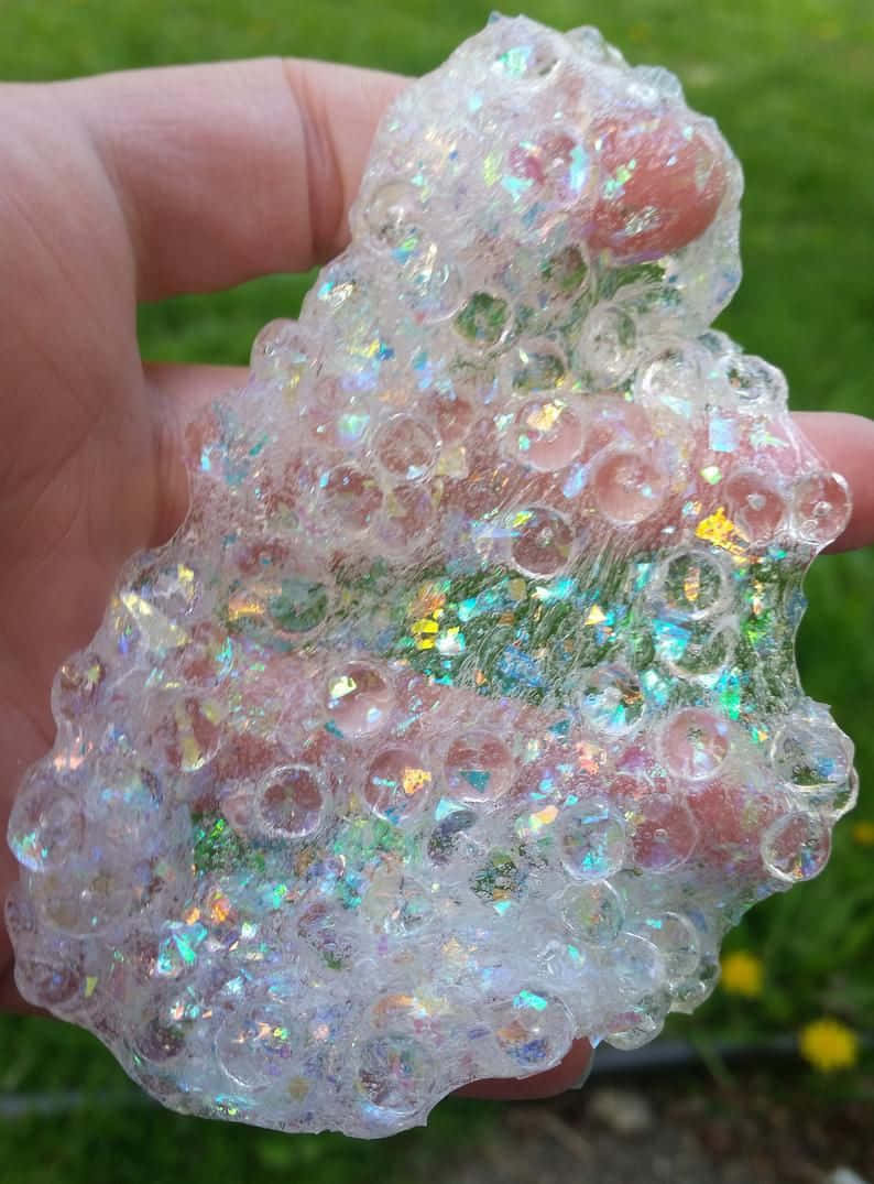slime 桌布 壁紙 — 一隻手拿著一個半透明、像泡泡糖一樣的史萊姆，裡面充滿了彩虹亮片和透明氣泡，背景是模糊的綠色草地。