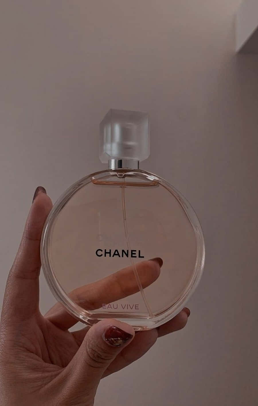 Chanel 桌布 壁紙 — 一隻手拿著一個圓形玻璃 Chanel Eau Vive 香水瓶，帶有磨砂蓋，靠著一面素牆。
