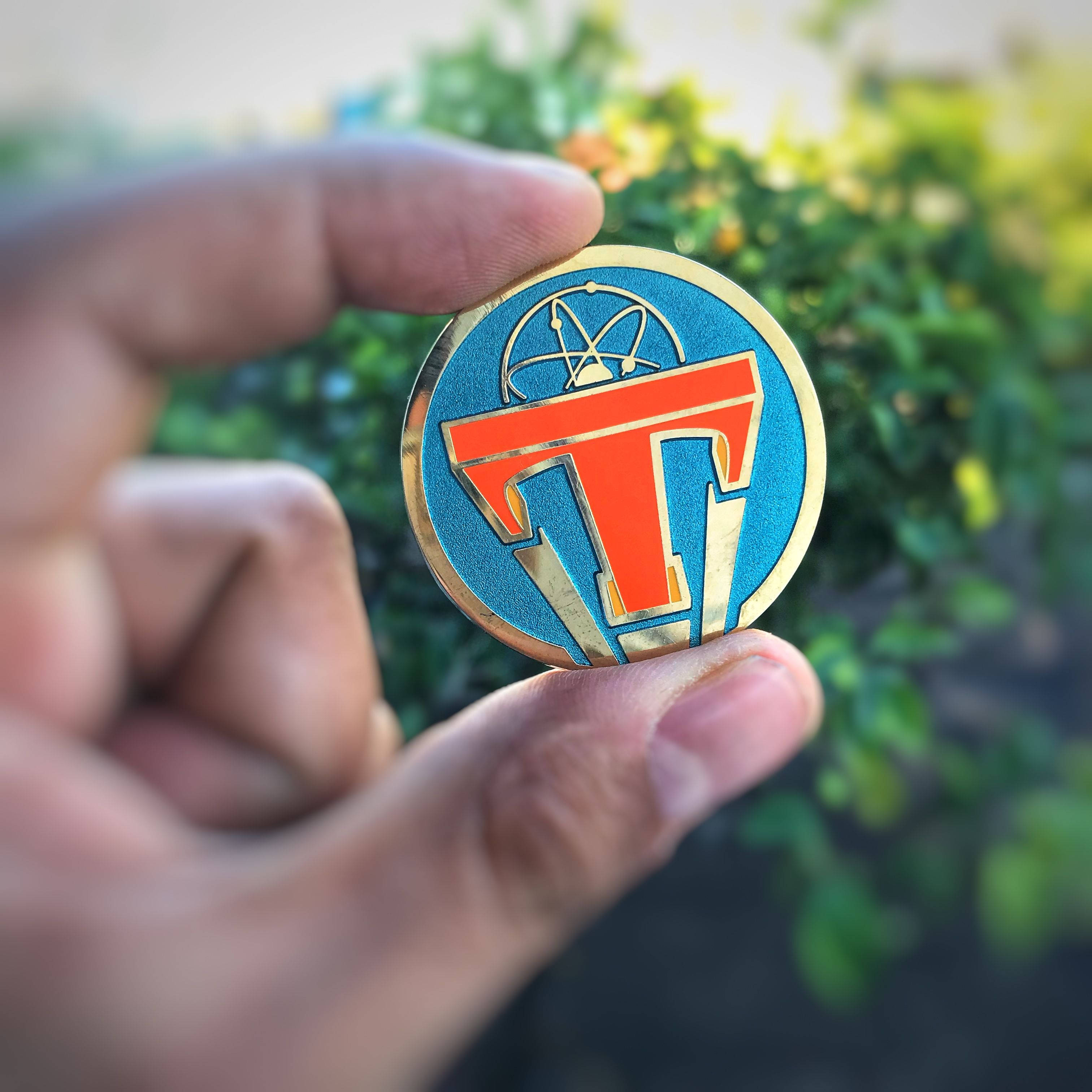明日世界電影 桌布 壁紙 — 一隻手拿著一個圓形琺瑯徽章，藍色背景上有大膽的橙色「T」和原子符號，背景是模糊的綠色樹葉。