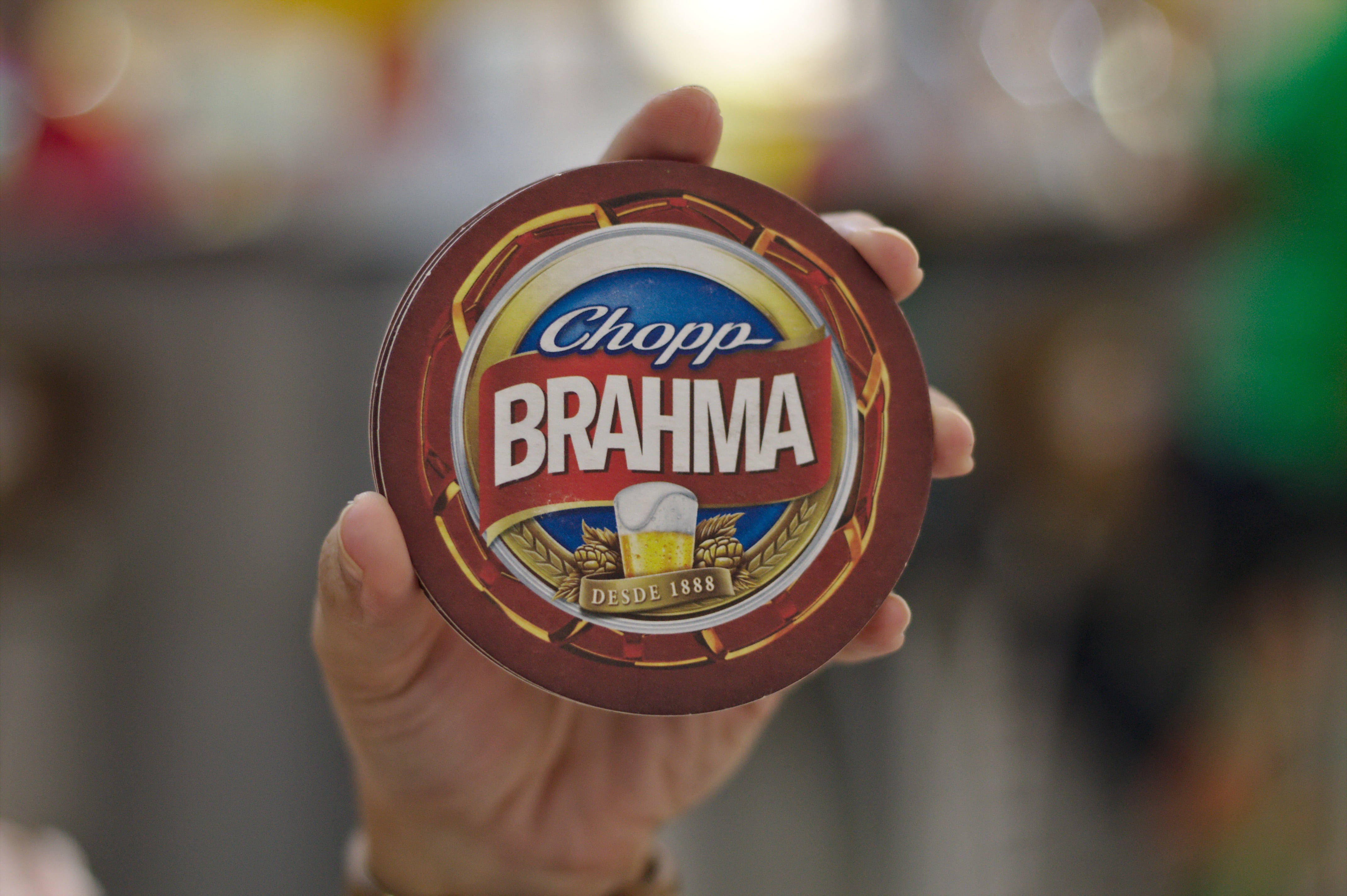 Beer 桌布 壁紙 — 一隻手拿著一個圓形的 Brahma 啤酒杯墊，上面有醒目的品牌標誌和金色小麥圖案，背景柔和模糊。