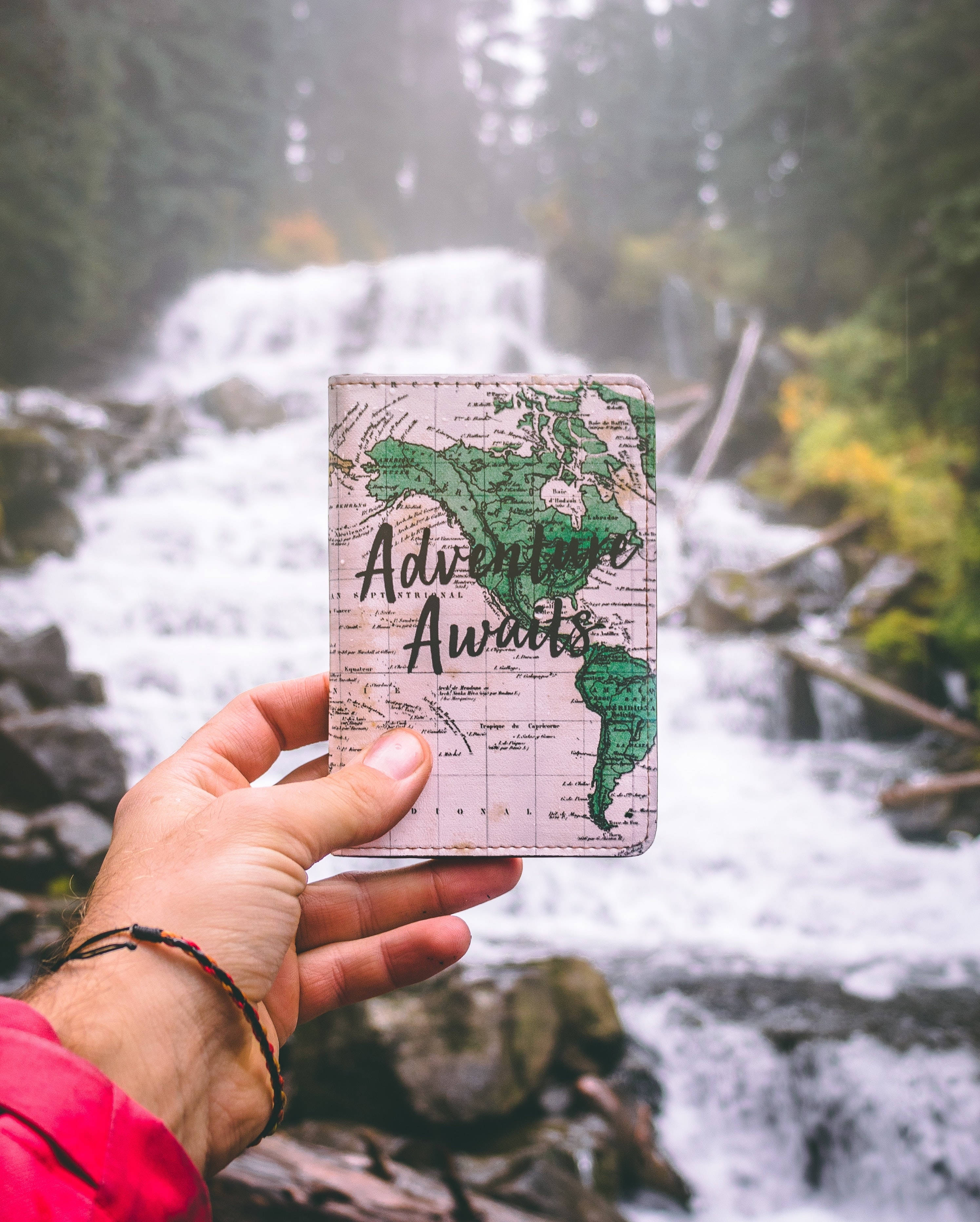 旅行 桌布 壁紙 — 一隻手拿著一個地圖主題的護照套，上面寫著「Adventure Awaits」，背景是霧濛濛的森林瀑布。