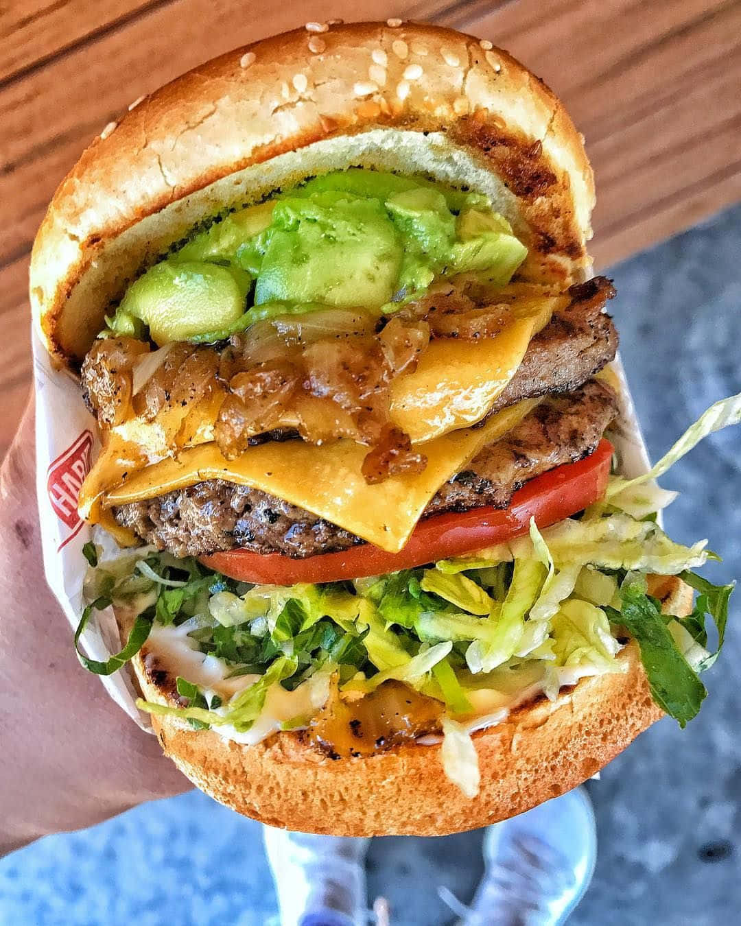 fast food 桌布 壁紙 — 一隻手拿著一個多汁的漢堡，裡面有融化的起司、酪梨、洋蔥、番茄和生菜，背景是木桌。