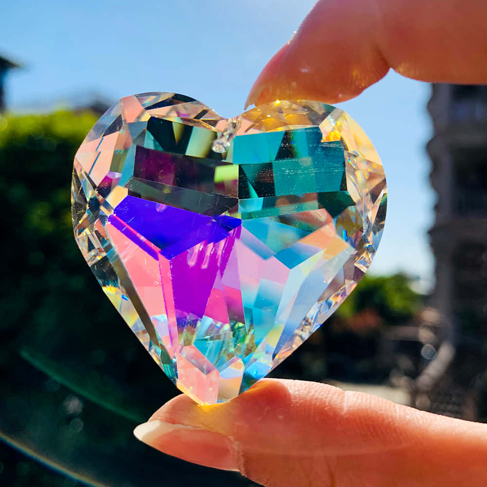 Gem Stone 桌布 壁紙 — 一隻手拿著一個多面切割的心形水晶稜鏡，在明亮的藍天下折射出鮮豔的彩虹色。