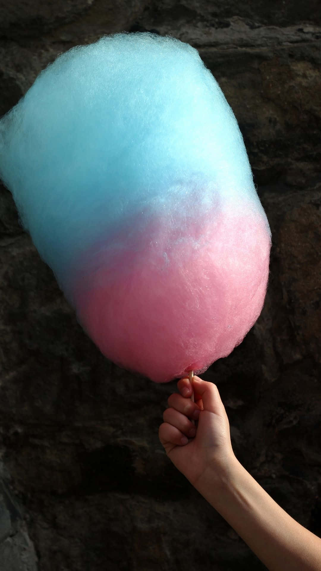 Cotton Candy 桌布 壁紙 — 一隻手拿著一個大棉花糖，帶有藍色到粉紅色的漸變，背景是深色石牆。