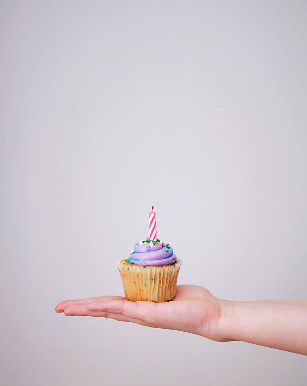 生日快樂 桌布 壁紙 — 一隻手拿著一個小杯子蛋糕，上面有紫色糖霜和粉紅色條紋蠟燭，背景是純灰色。