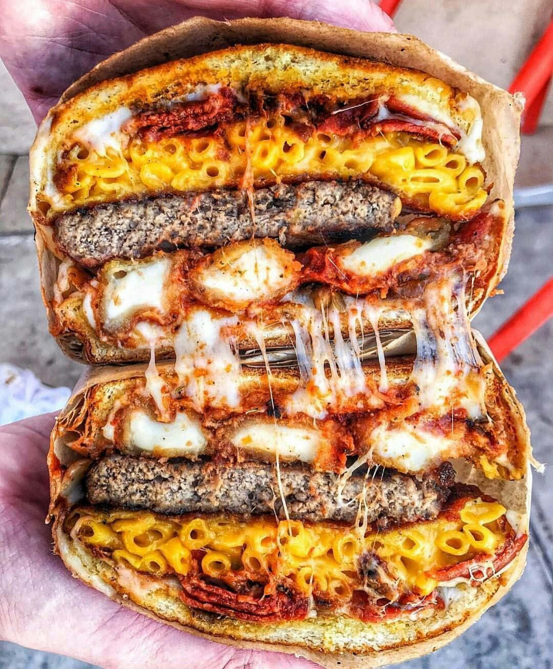 fast food 桌布 壁紙 — 一隻手拿著一個巨型漢堡，切成兩半，露出通心粉起司、肉餅、炸雞和融化起司的層次。