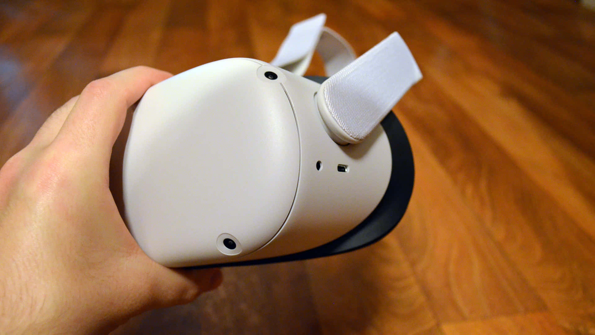 Oculus Quest 2 桌布 壁紙 — 一隻手拿著一個帶有可調節頭帶的白色VR頭戴式顯示器，背景是溫暖的木地板。