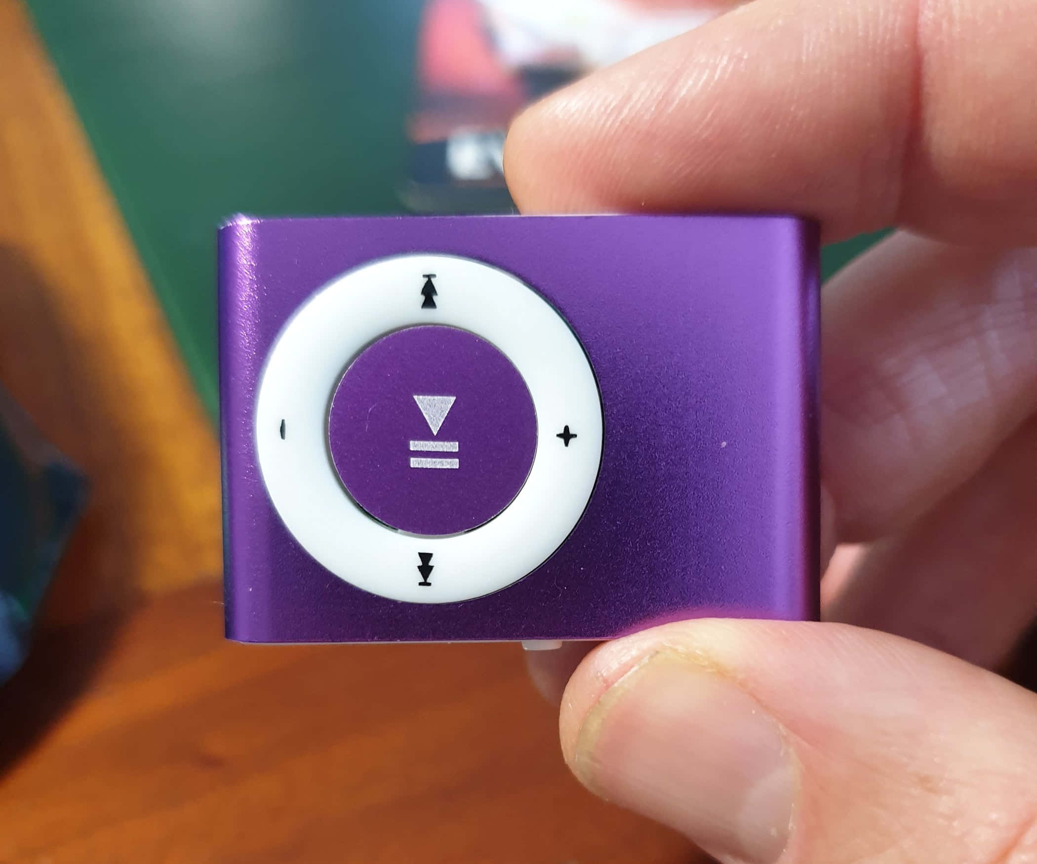 Music Player 桌布 壁紙 — 一隻手拿著一個帶有白色點擊輪的紫色 iPod shuffle，背景是模糊的木紋。
