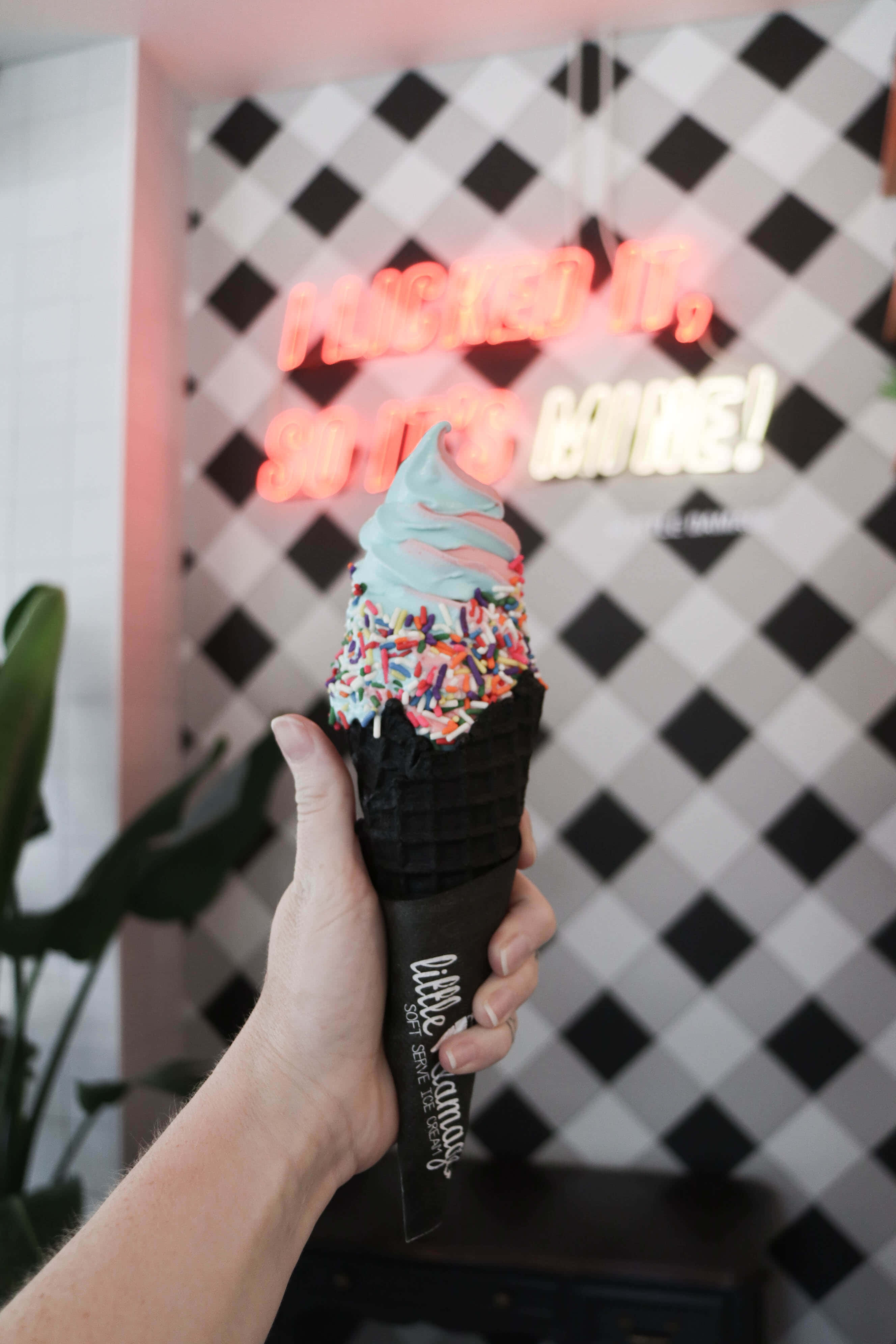 Ice Cream 桌布 壁紙 — 一隻手拿著一個帶有粉彩漩渦和彩色糖粒的軟冰淇淋甜筒，背景是黑白格紋牆，上面有發光的霓虹燈文字。
