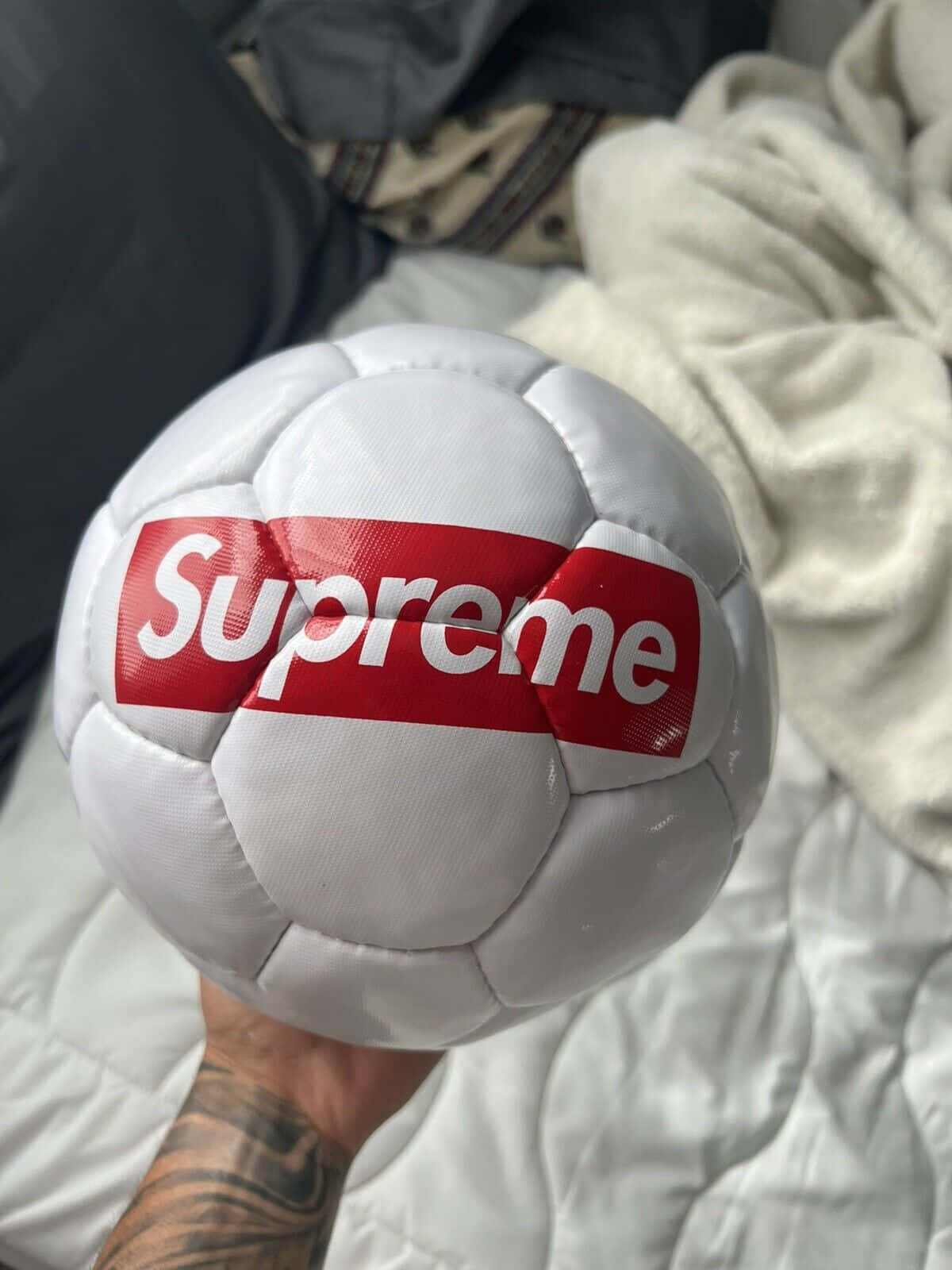 supreme 桌布 壁紙 — 一隻手拿著一個帶有紅色Supreme標誌的白色足球，背景是皺巴巴的白色毯子和深色衣服。