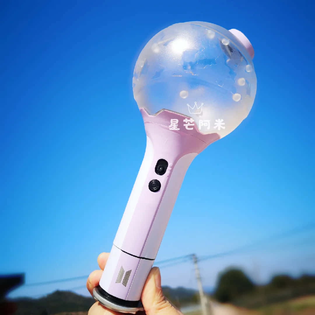 Bts Army Bomb 桌布 壁紙 — 一隻手拿著一個帶有透明泡泡頂部的粉紅色發光棒，背景是明亮的藍天。