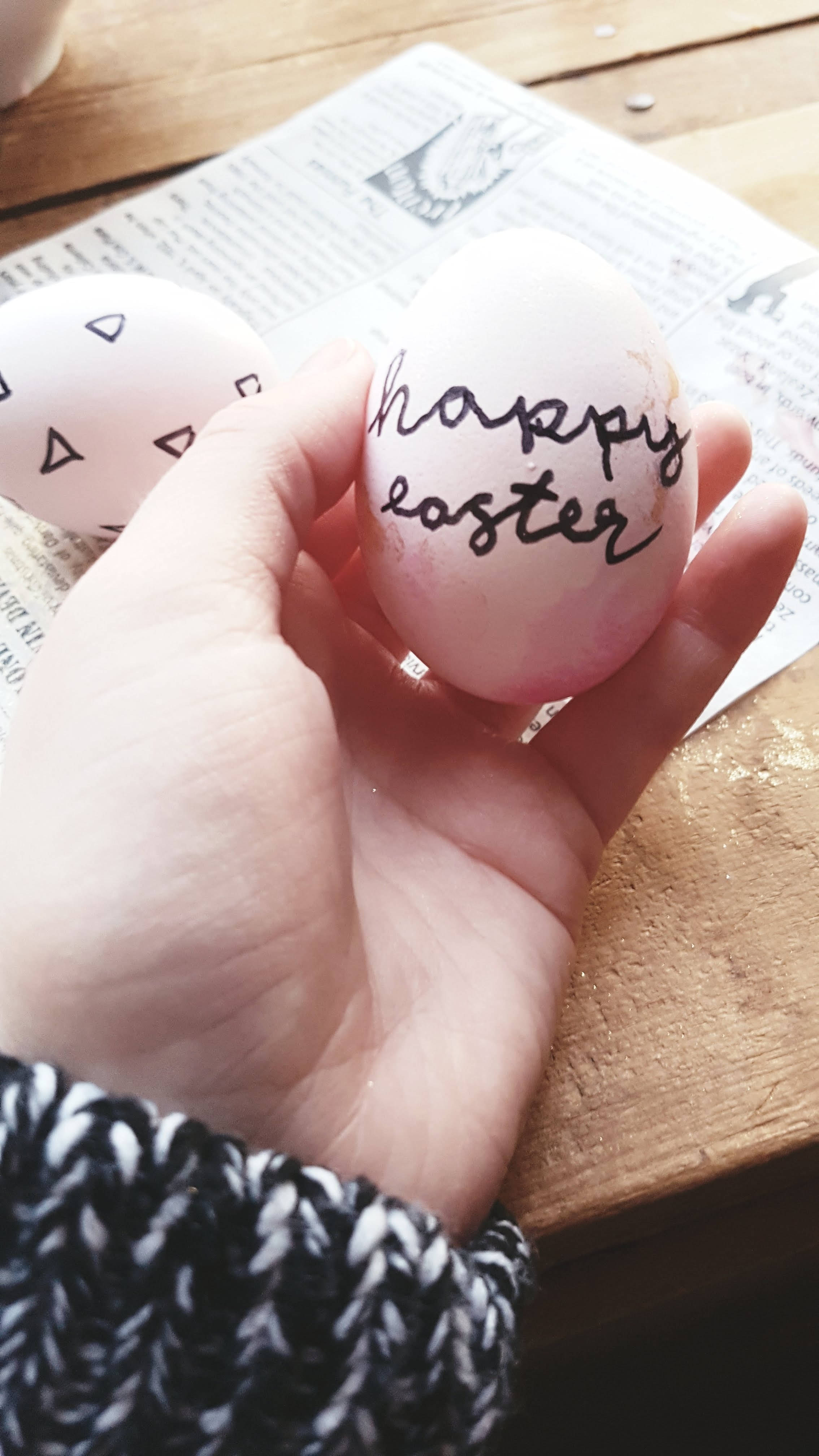 Happy Easter 桌布 壁紙 — 一隻手拿著一個手繪復活蛋，上面有草書 'happy easter'，放在鋪有報紙的木桌上。