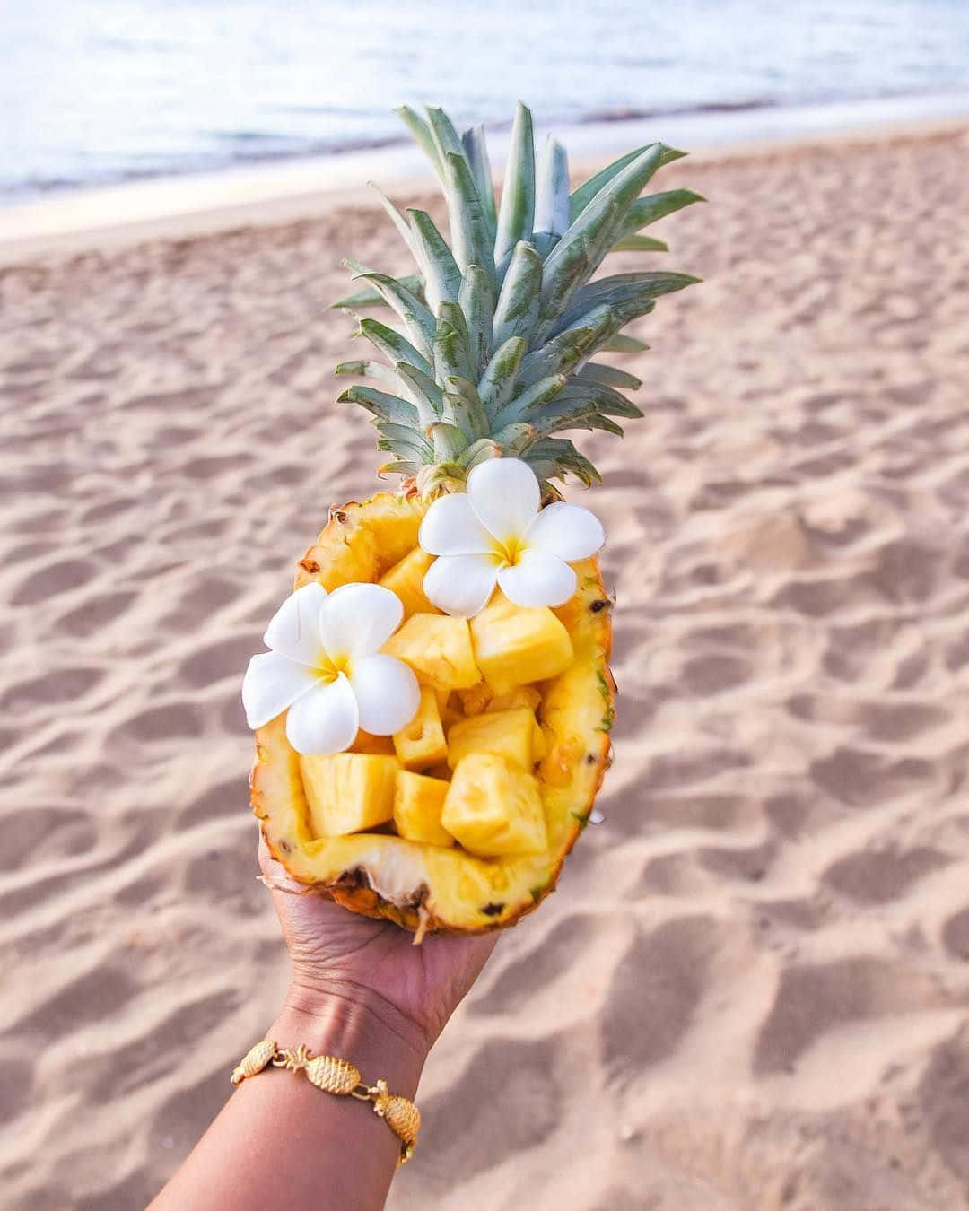 Pineapple 桌布 壁紙 — 一隻手拿著一個挖空的鳳梨，裡面裝滿多汁的果肉和白色雞蛋花，背景是沙灘。