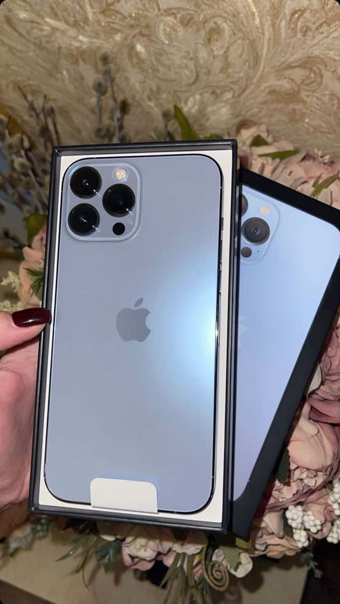 iPhone 12 桌布 壁紙 — 一隻手拿著一個新的淺藍色 iPhone 13 Pro 放在盒子裡，周圍環繞著柔和的粉紅色花朵和華麗的壁紙。