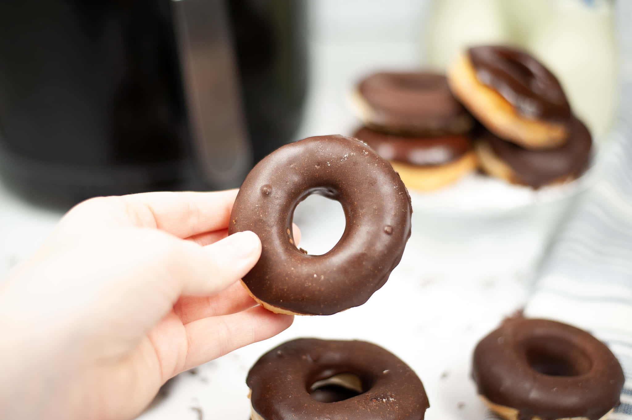Glazed Donut 桌布 壁紙 — 一隻手拿著一個有光澤的巧克力釉面甜甜圈，其他甜甜圈散落在附近的白色表面上。